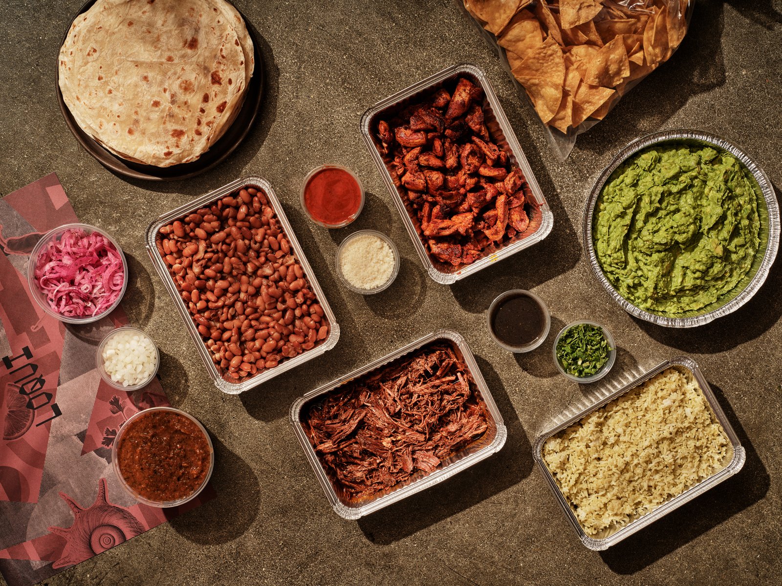Family-style catering trays of chicken, shredded meat, beans, and rice with tortillas, chips, and guacamole