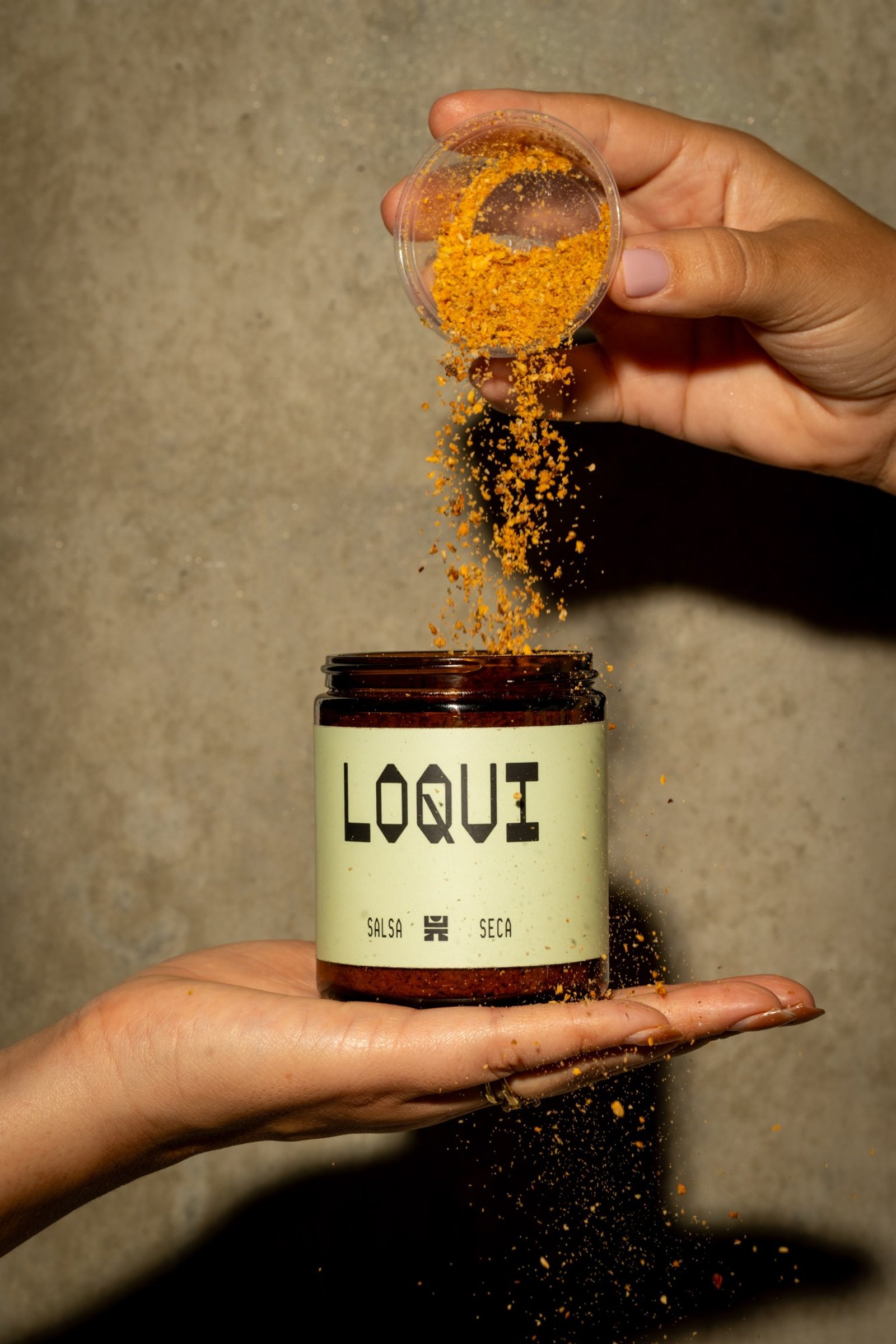 Hands sprinkling seasoning into a jar of LOQUI salsa seca