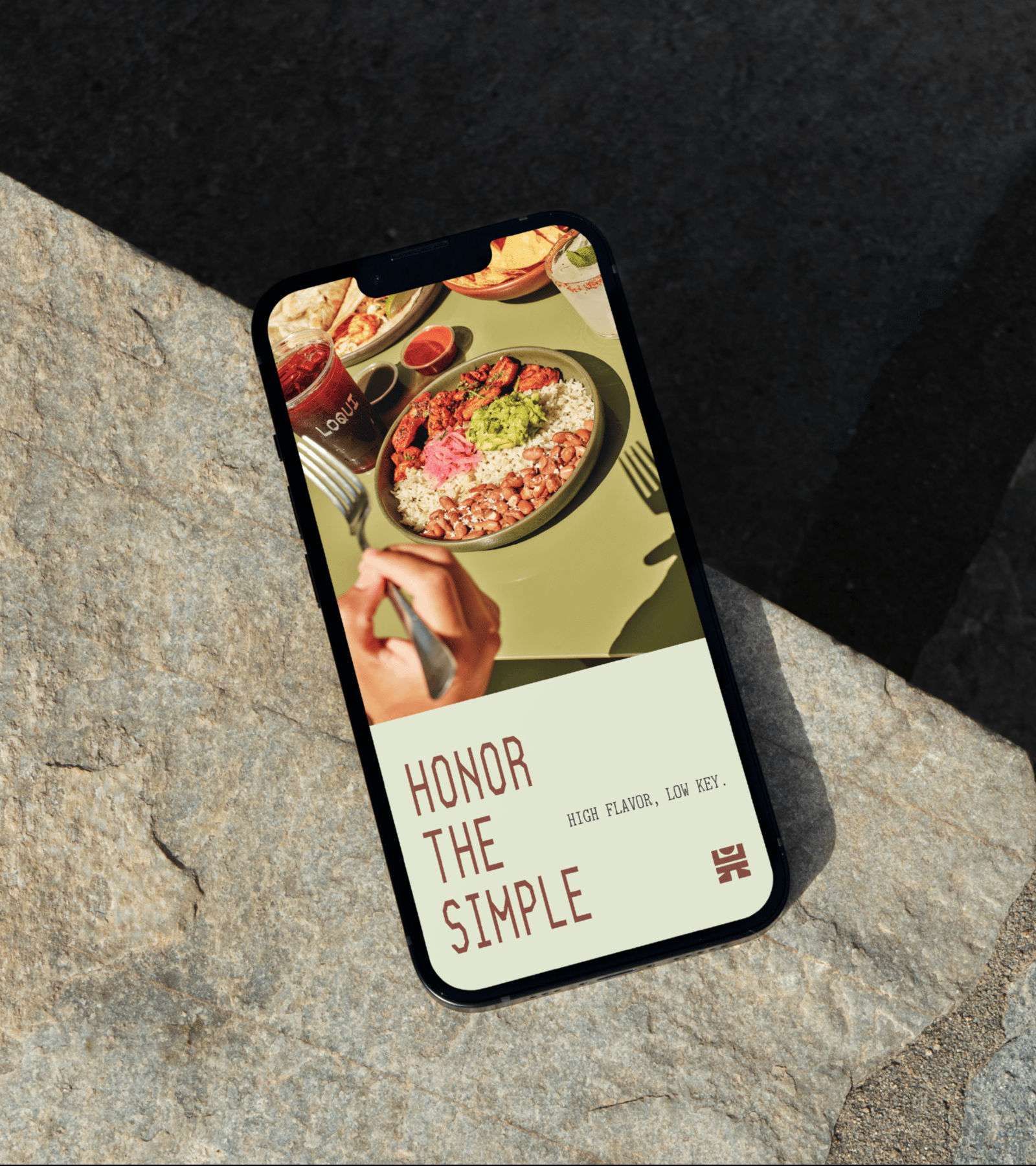 Phone displaying LOQUI food and the words Honor the Simple on a sunlit stone surface