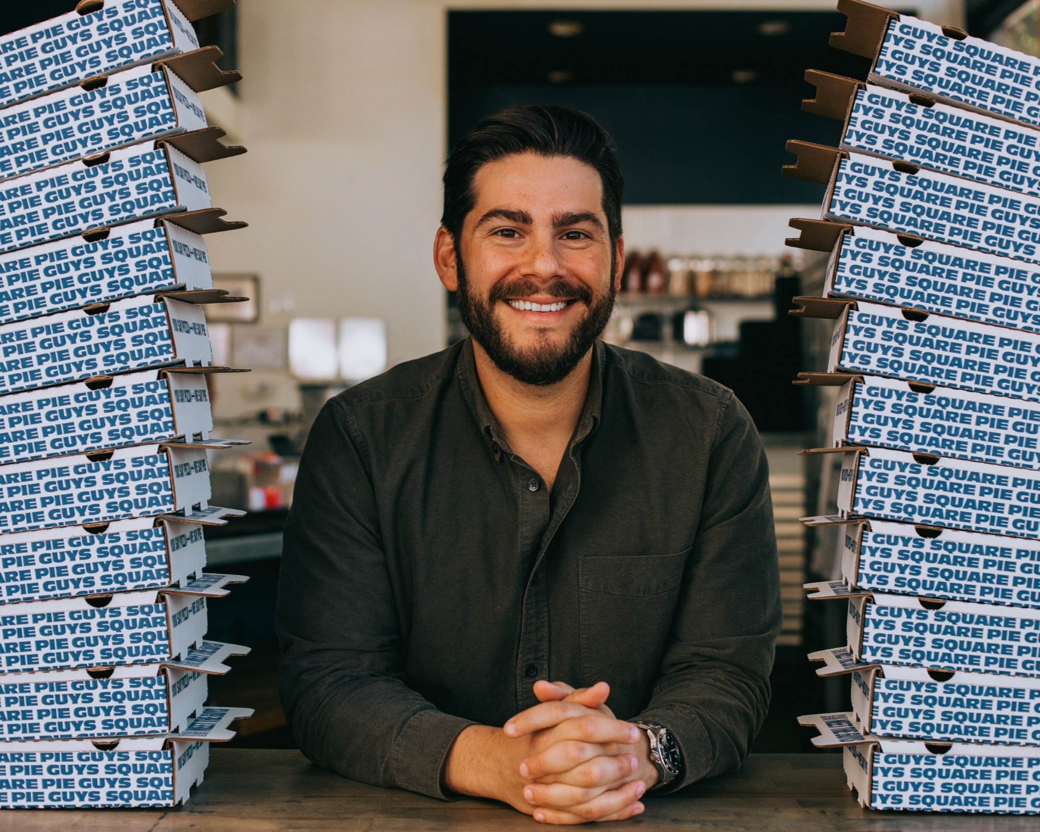 Marc Schechter, founder of Square Pie Guys and author of Square Pie Guy: Detroit-Style Pizza at Home, smiling between two stacks of Square Pie Guys pizza boxes.