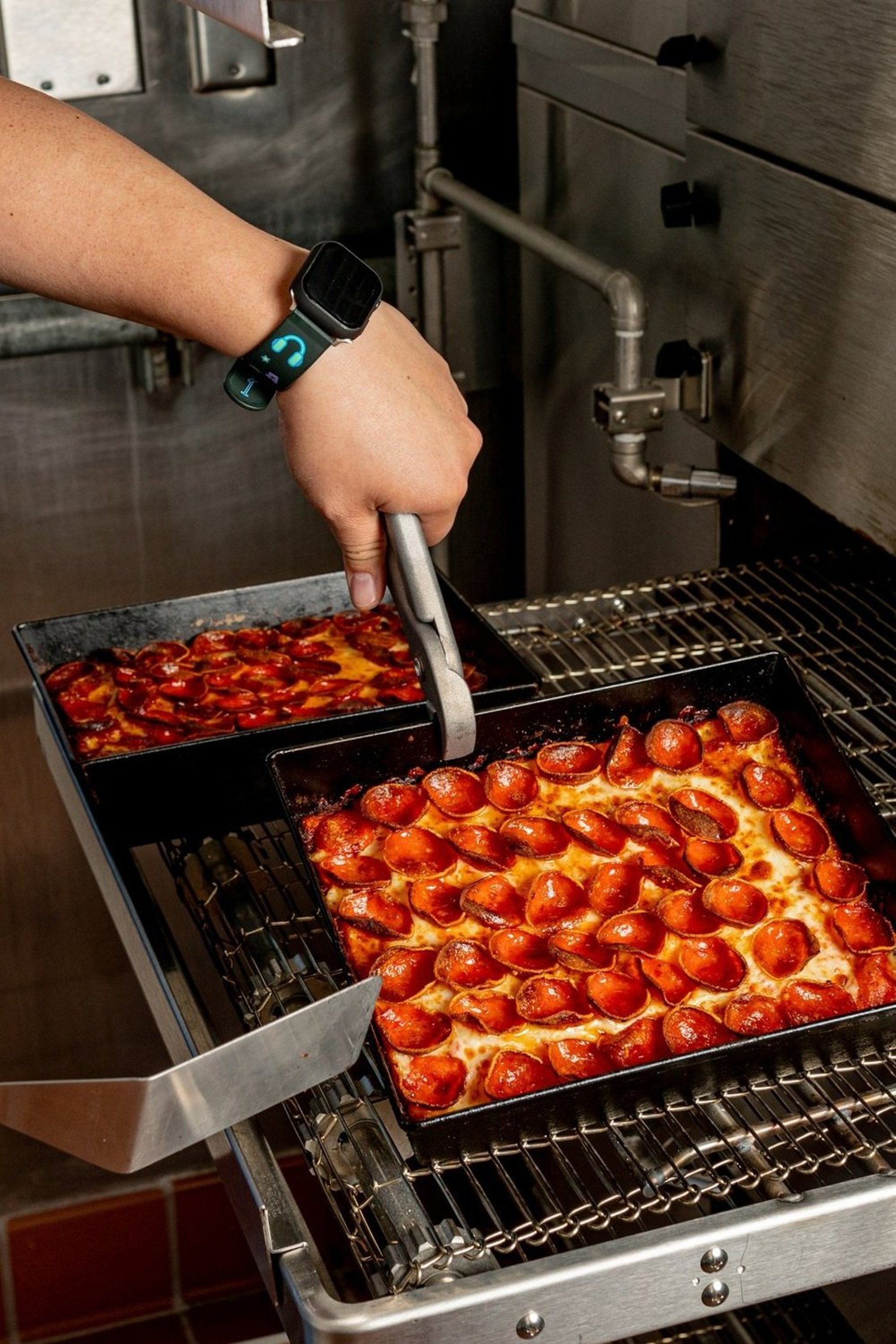 A person is placing freshly baked pepperoni pizzas into an oven using tongs. The person is wearing a smartwatch on their wrist.