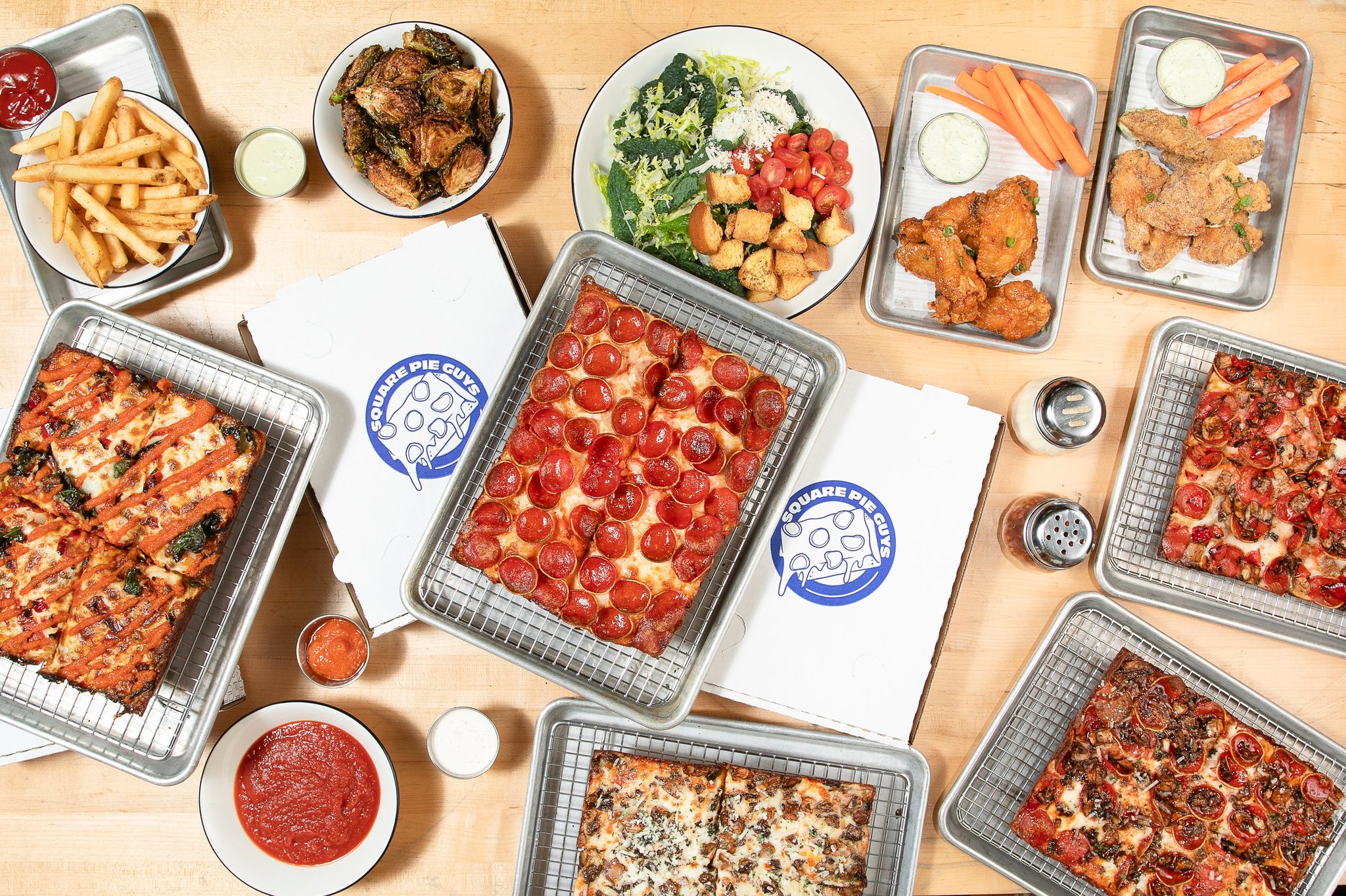 Four trays of different varieties of rectangular pizza on a wooden table.