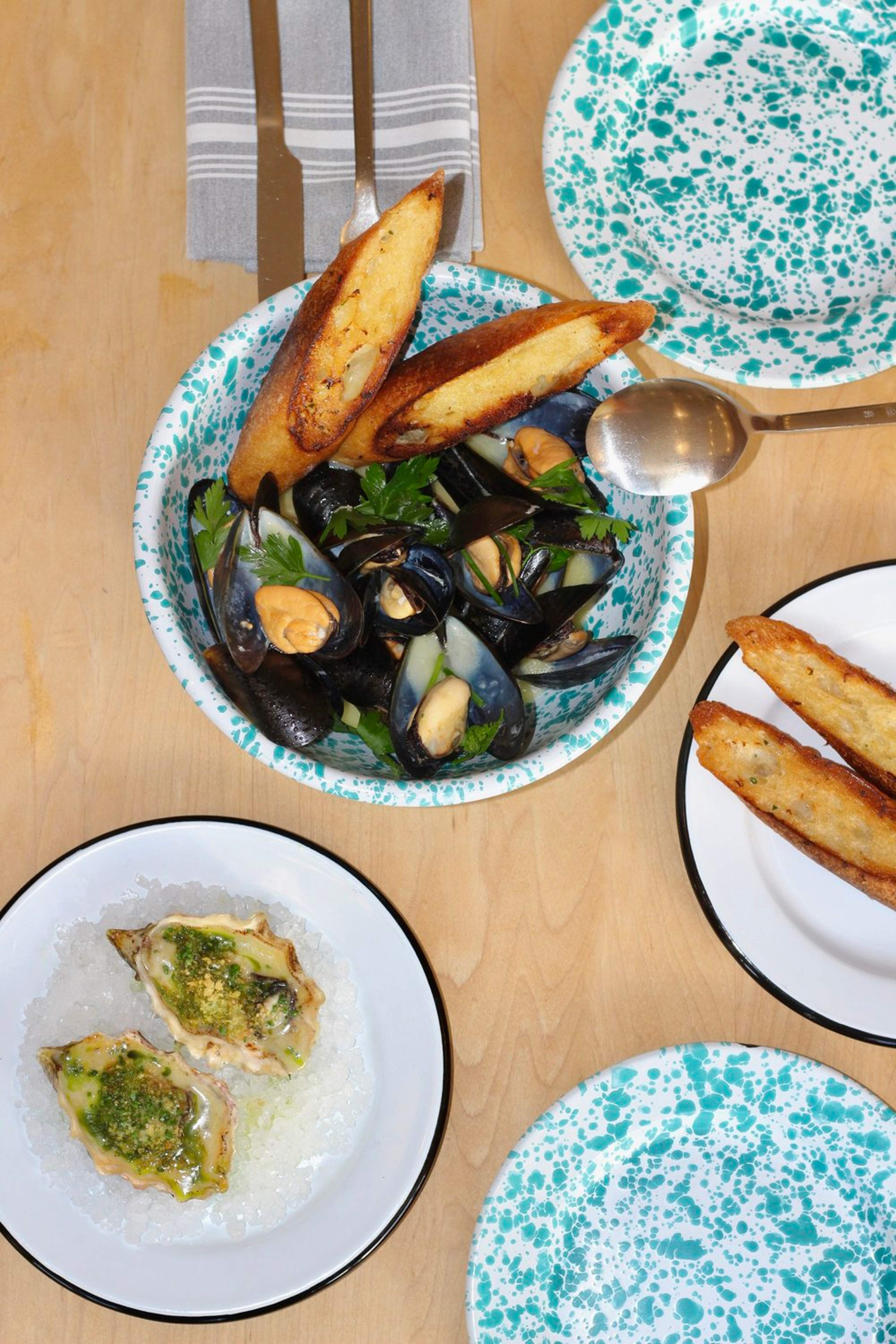 A table with a dish of cooked mussels garnished with green herbs, two toasted bread slices, and a plate of oysters topped with green sauce.