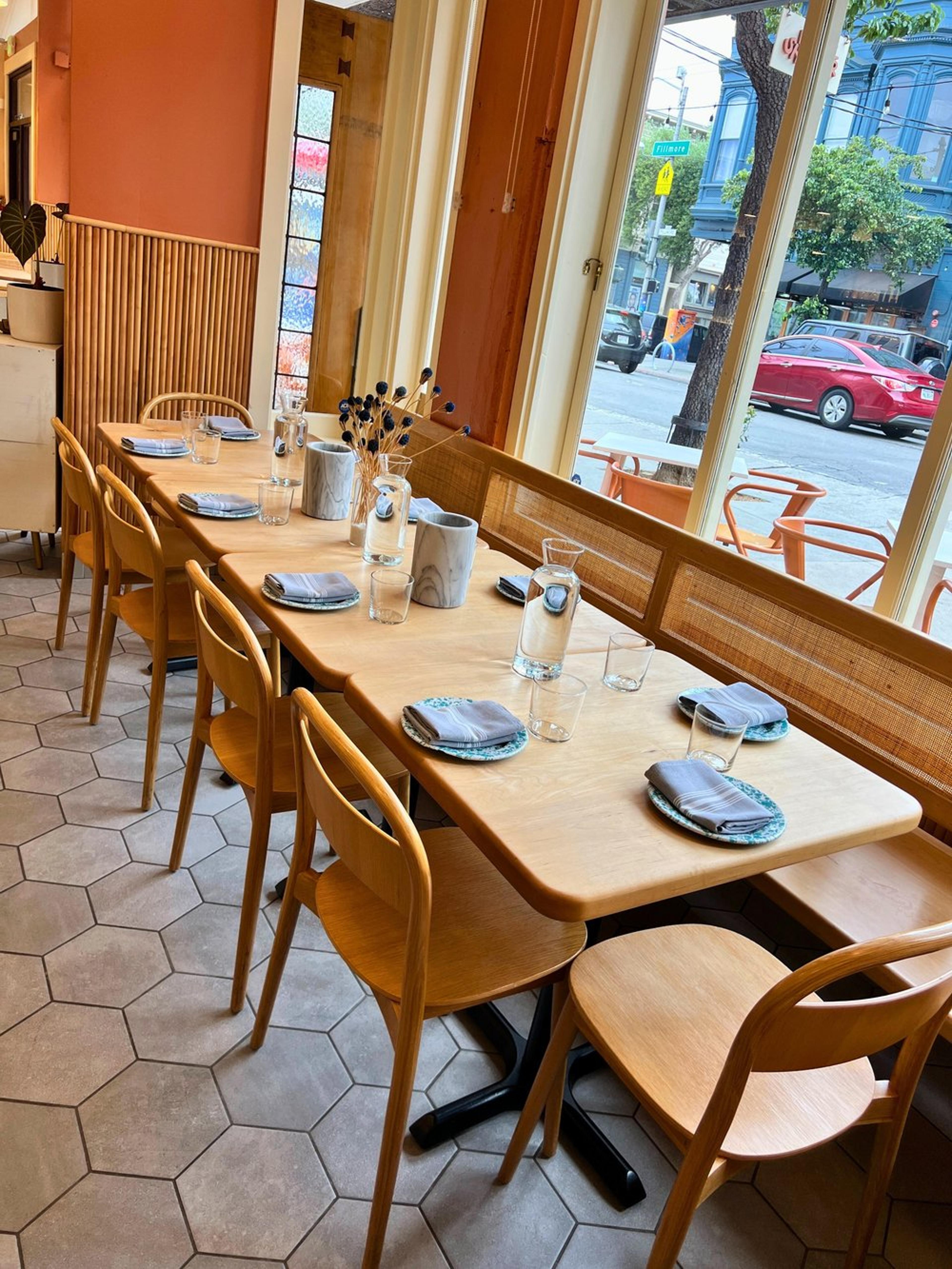 A cozy restaurant interior with a long wooden table set for dining, including glasses, napkins, and decorative vases with dried flowers, next to large windows showing a city street with parked cars and buildings outside.