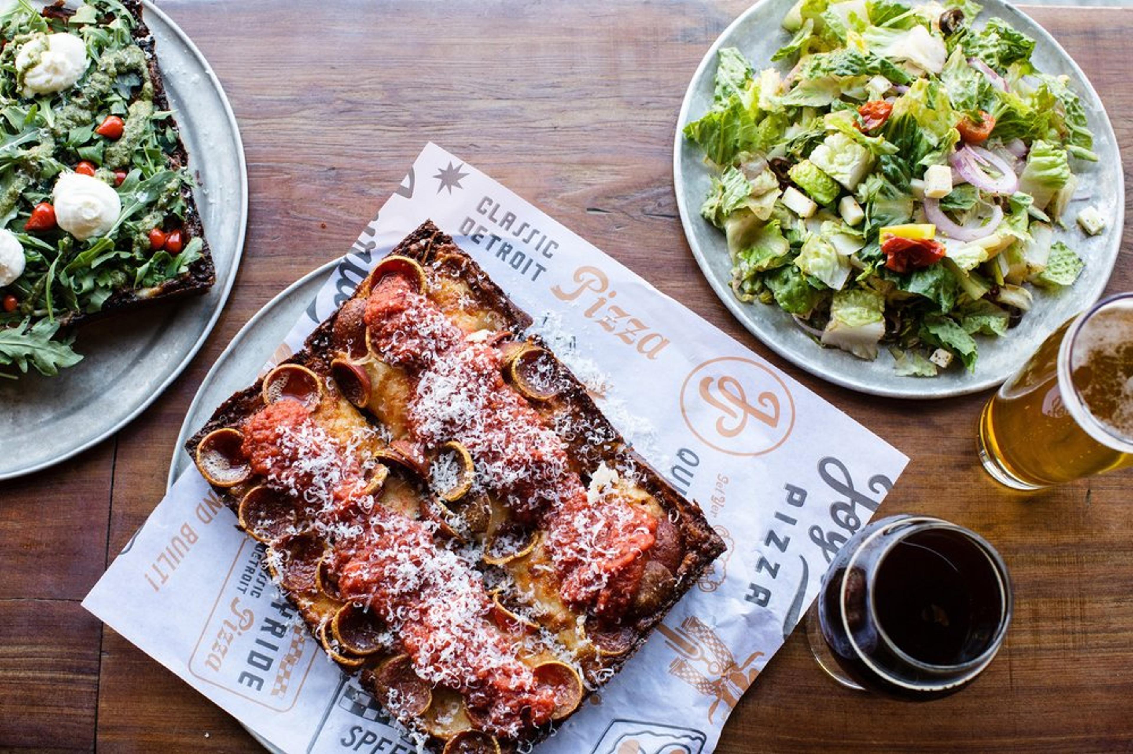 A top-down view of a meal featuring a rectangular Detroit-style pepperoni pizza on branded paper. Next to it is a green salad with sliced vegetables, a glass of beer, a soda, and a pizza topped with arugula and cherry tomatoes.