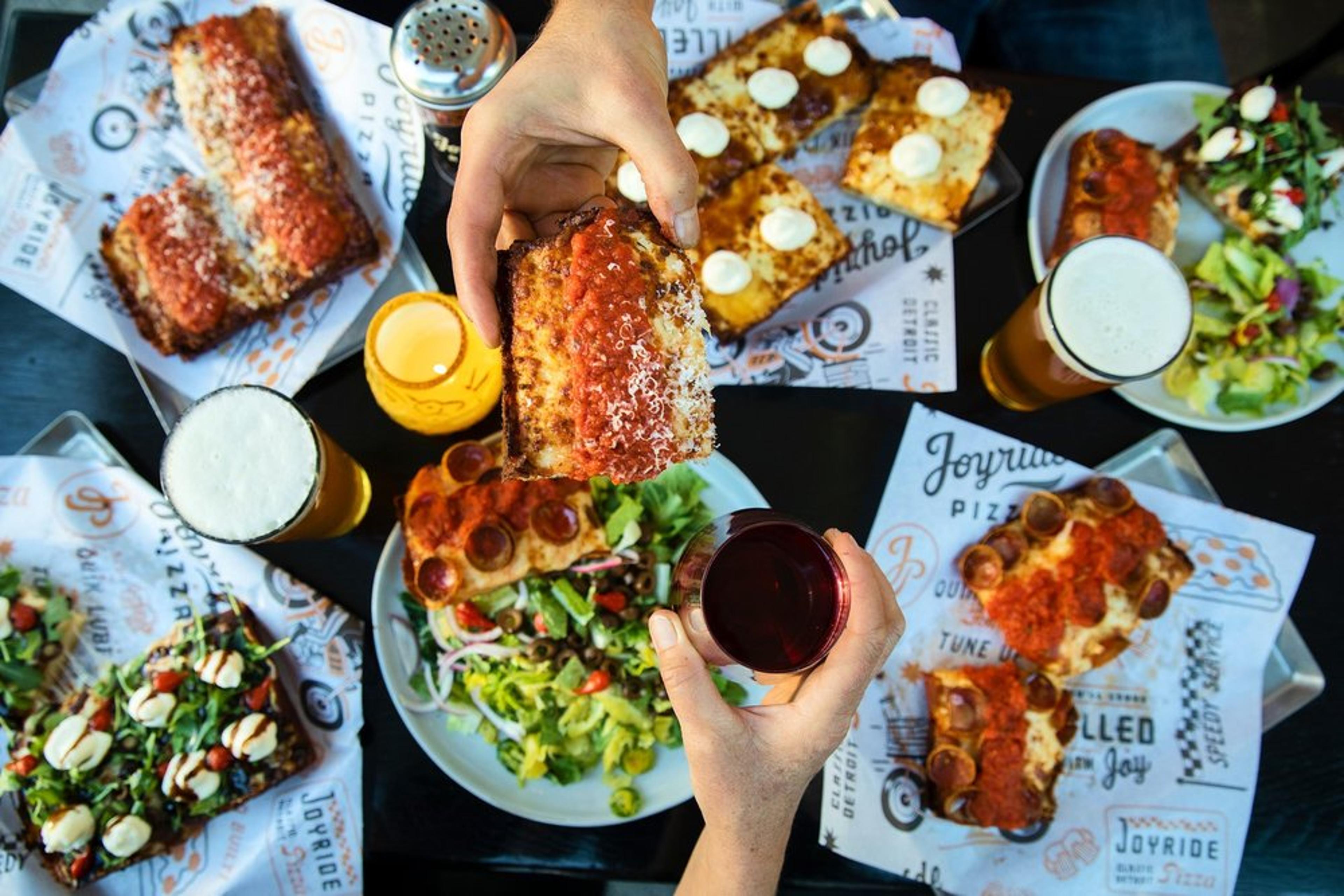 Hands holding a slice of pizza and a glass of red wine over a table filled with various pizzas, salads, and drinks. Several plates and glasses are visible, along with paper liners featuring a restaurant logo.