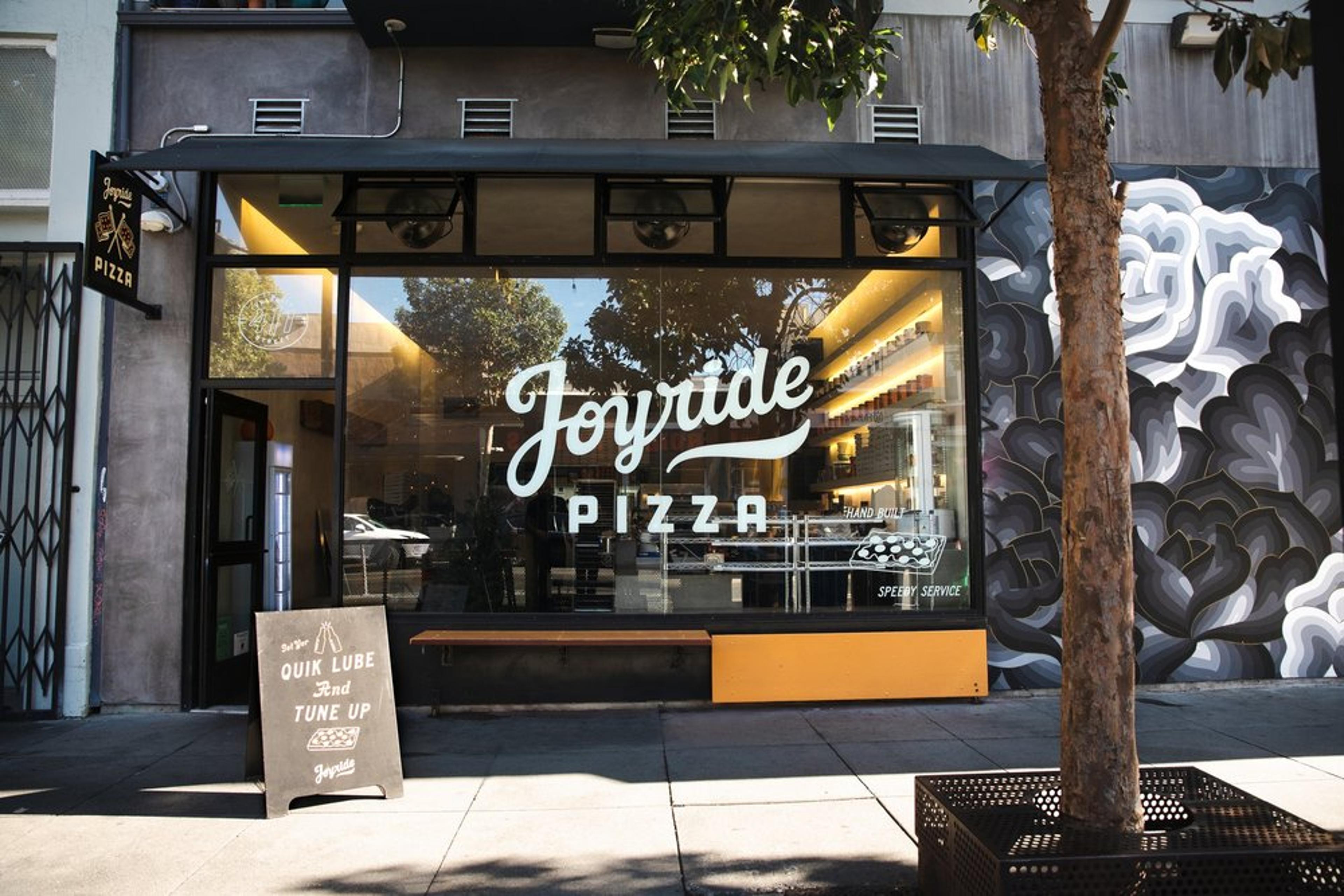 Street view of Joyride Pizza with a glass storefront featuring the logo in white. A chalkboard sign outside reads Oil, Lube and Tune Up. A tree partially shades the entrance, and a mural with floral patterns decorates the adjacent wall.