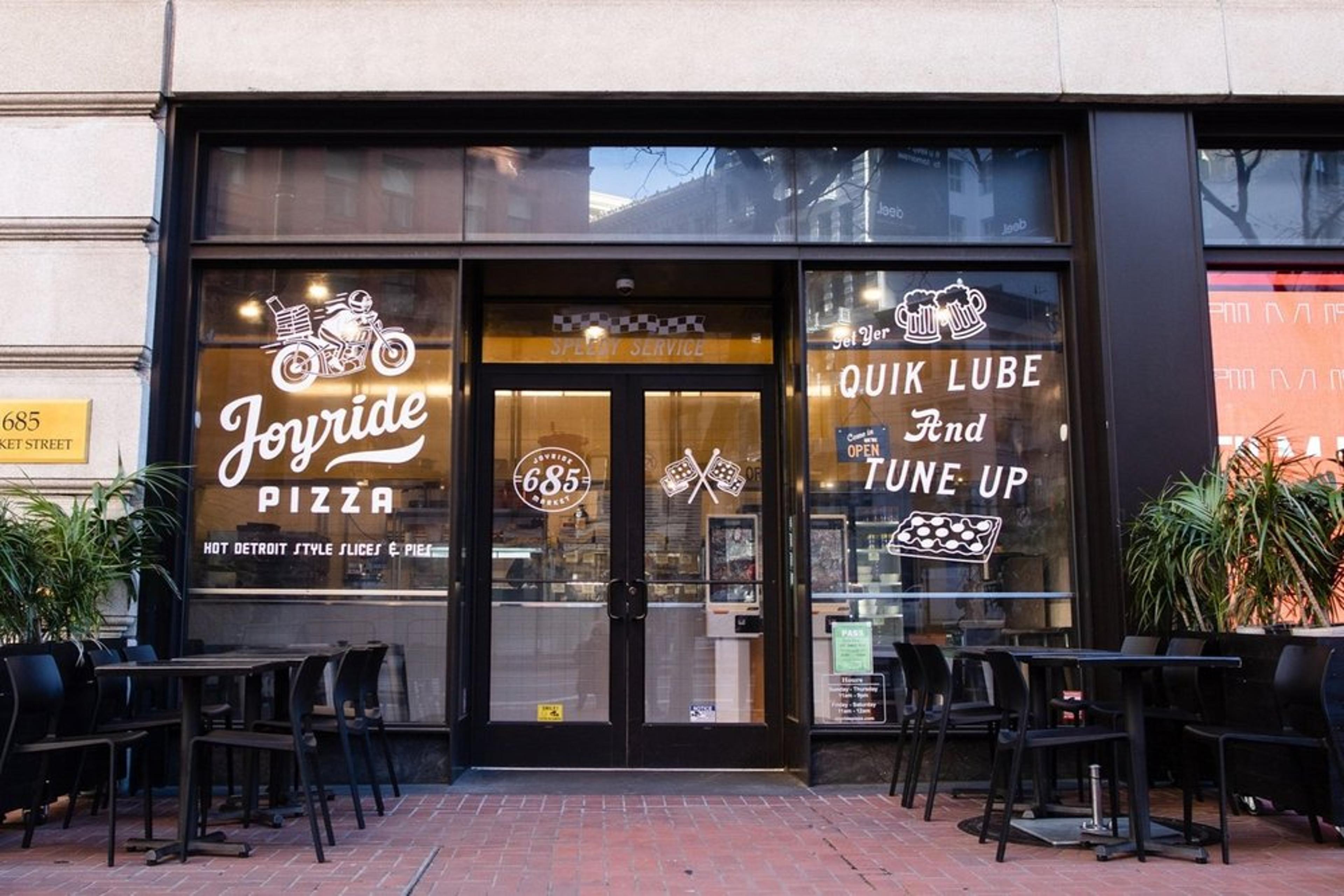 Street view of Joyride Pizza, featuring large glass windows with white graphic designs and text promoting pizza, drinks, and car-themed décor. Black stools and tables are arranged on the brick sidewalk outside.