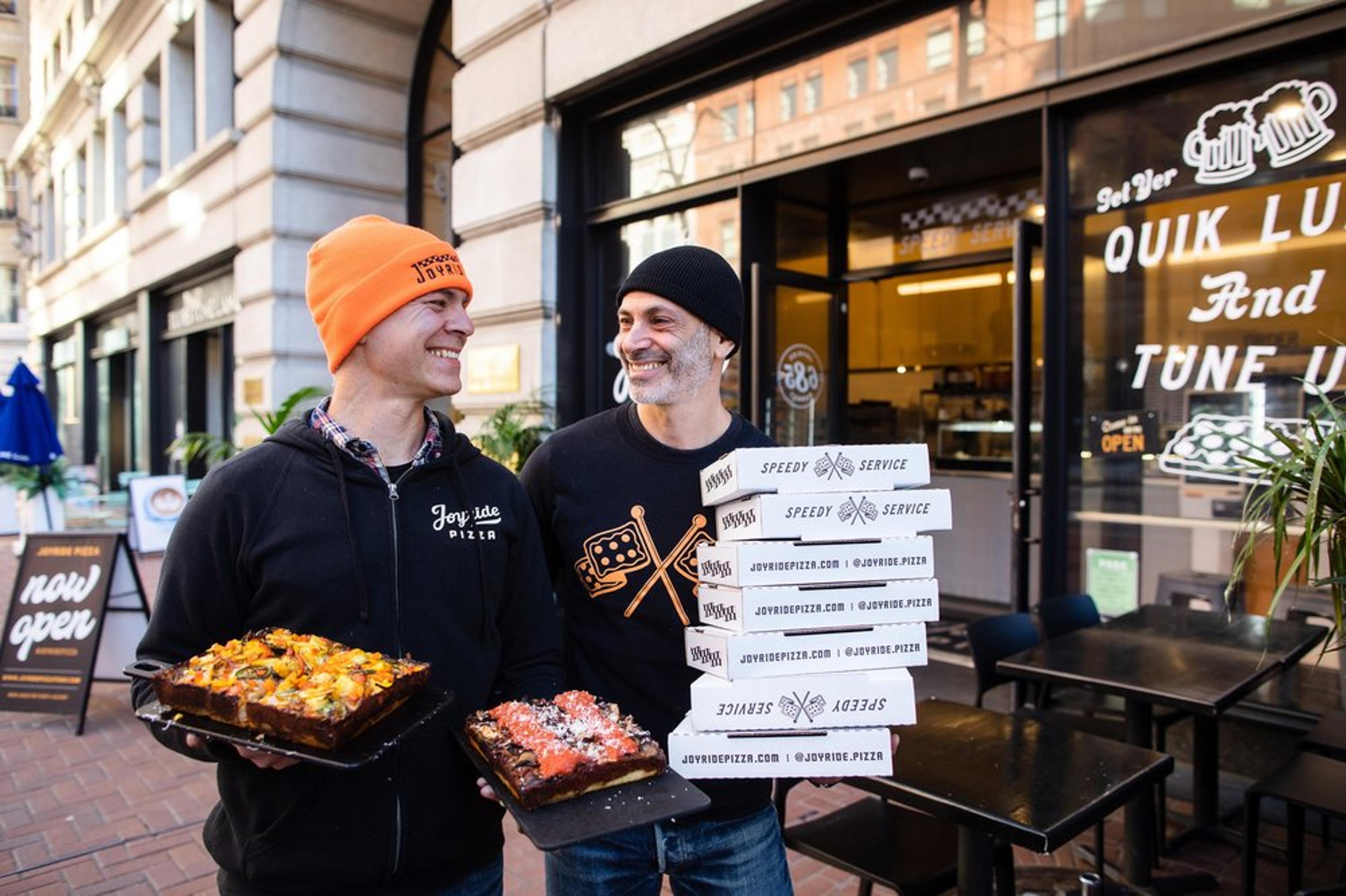 Two men stand outside a pizzeria. One holds two pans of pizza, while the other carries a stack of pizza boxes. They are smiling, wearing hats and casual clothing, with the restaurant's storefront and outdoor seating visible in the background.