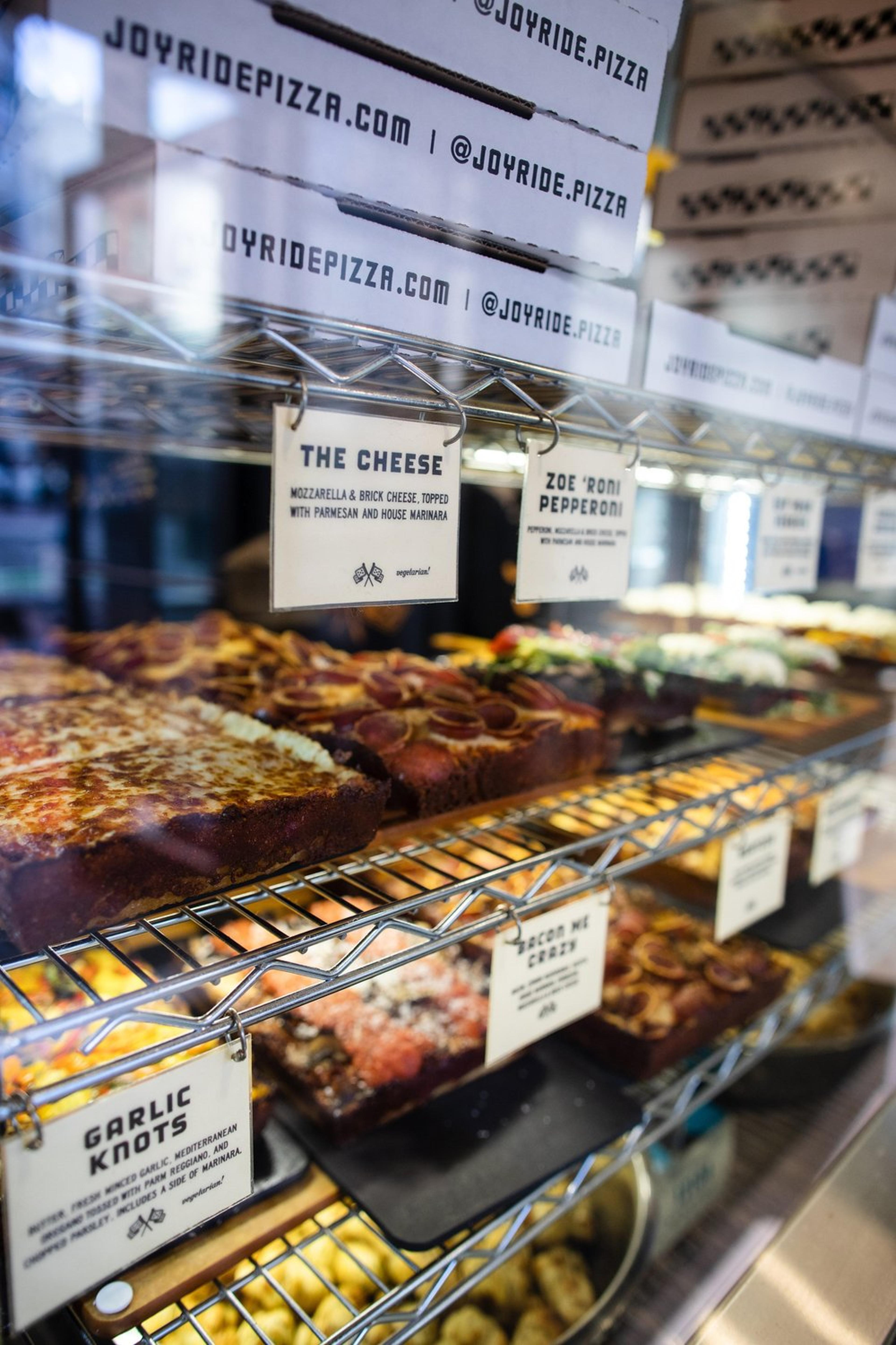 Pizza and garlic knots on display in a restaurant window. Various pizza flavors such as The Cheese and Zoe Roni Pepperoni are labeled. Pizza boxes are stacked above, and restaurant social media handles are visible on the window.