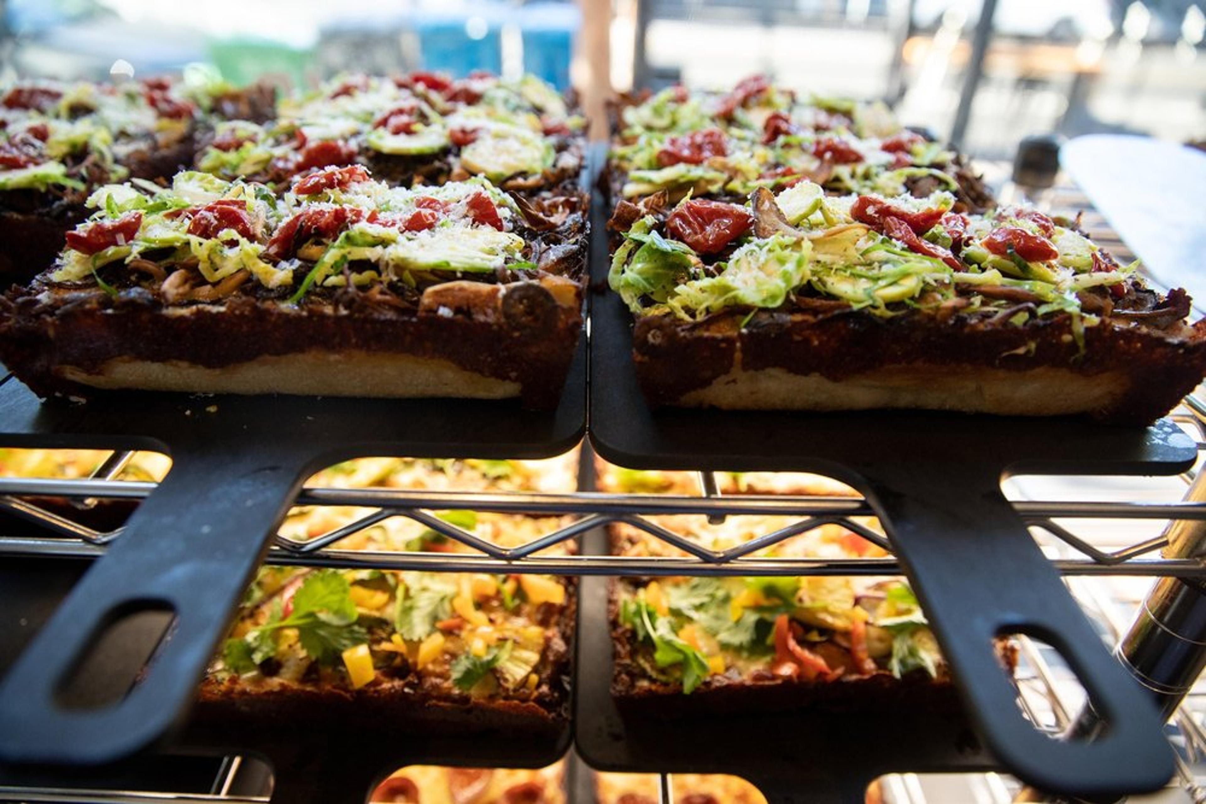 Slices of thick-crust pizza topped with shredded lettuce, tomatoes, and cheese displayed on black trays. The pizzas have a variety of colorful toppings and are set on metal racks in a well-lit setting.