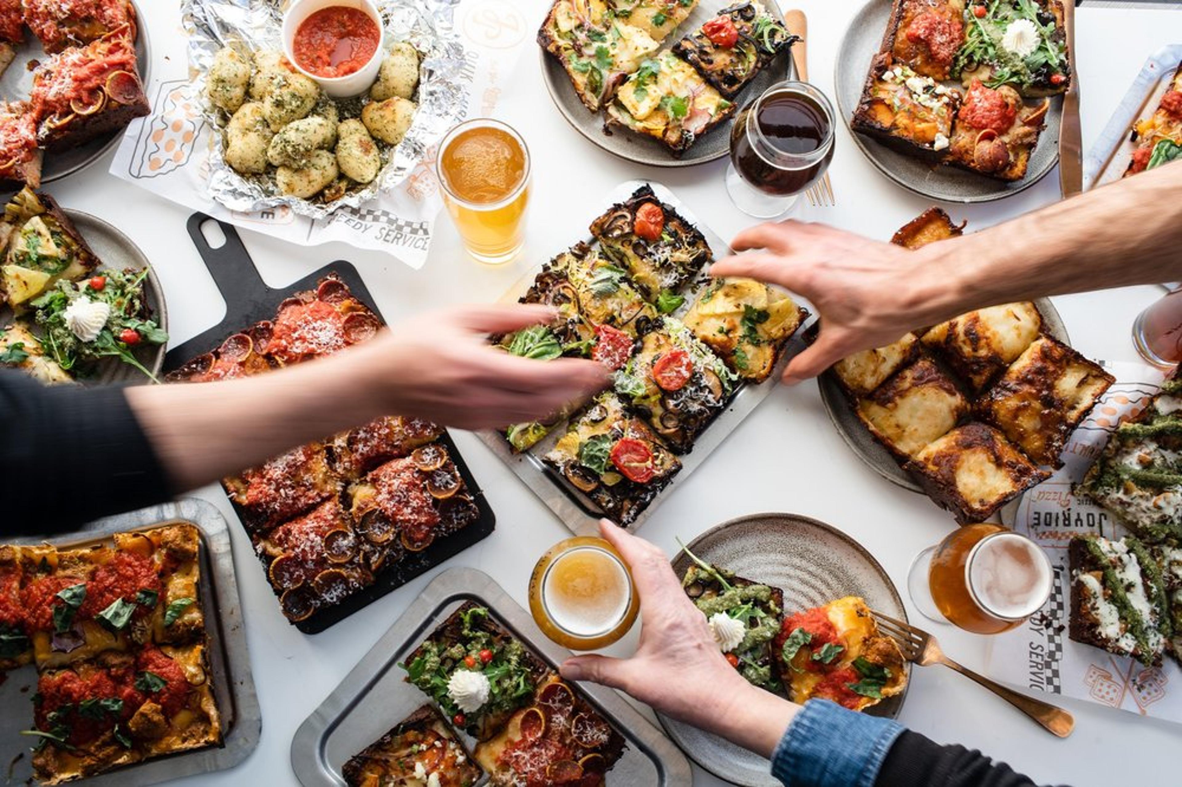 A spread of Joyride Detroit-style pizzas with people reaching for slices