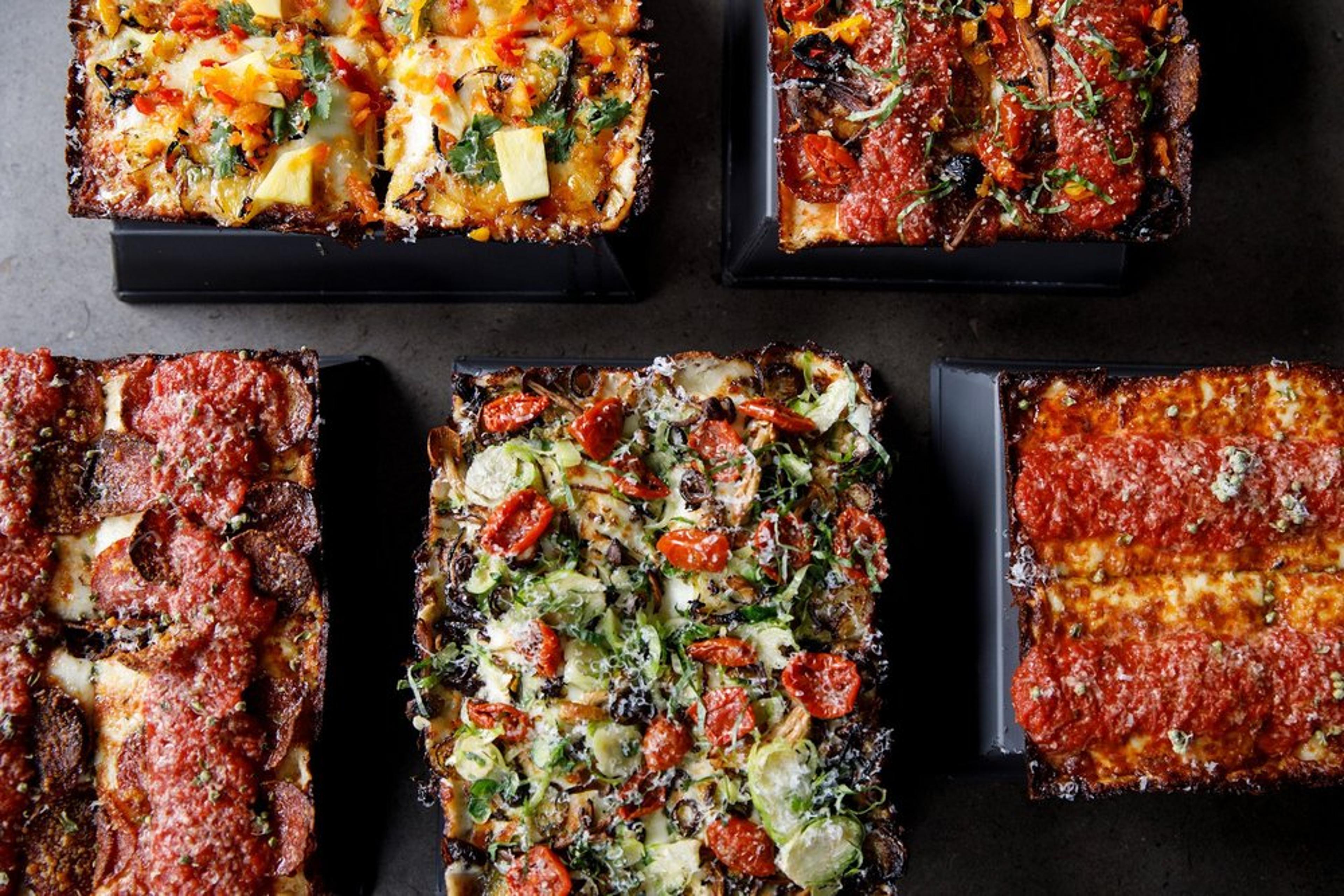 A top view of various rectangular Detroit-style pizzas on dark trays. Each pizza is topped with different ingredients, including pepperoni, cherry tomatoes, mushrooms, and herbs, highlighting their crispy, cheese-laden edges.