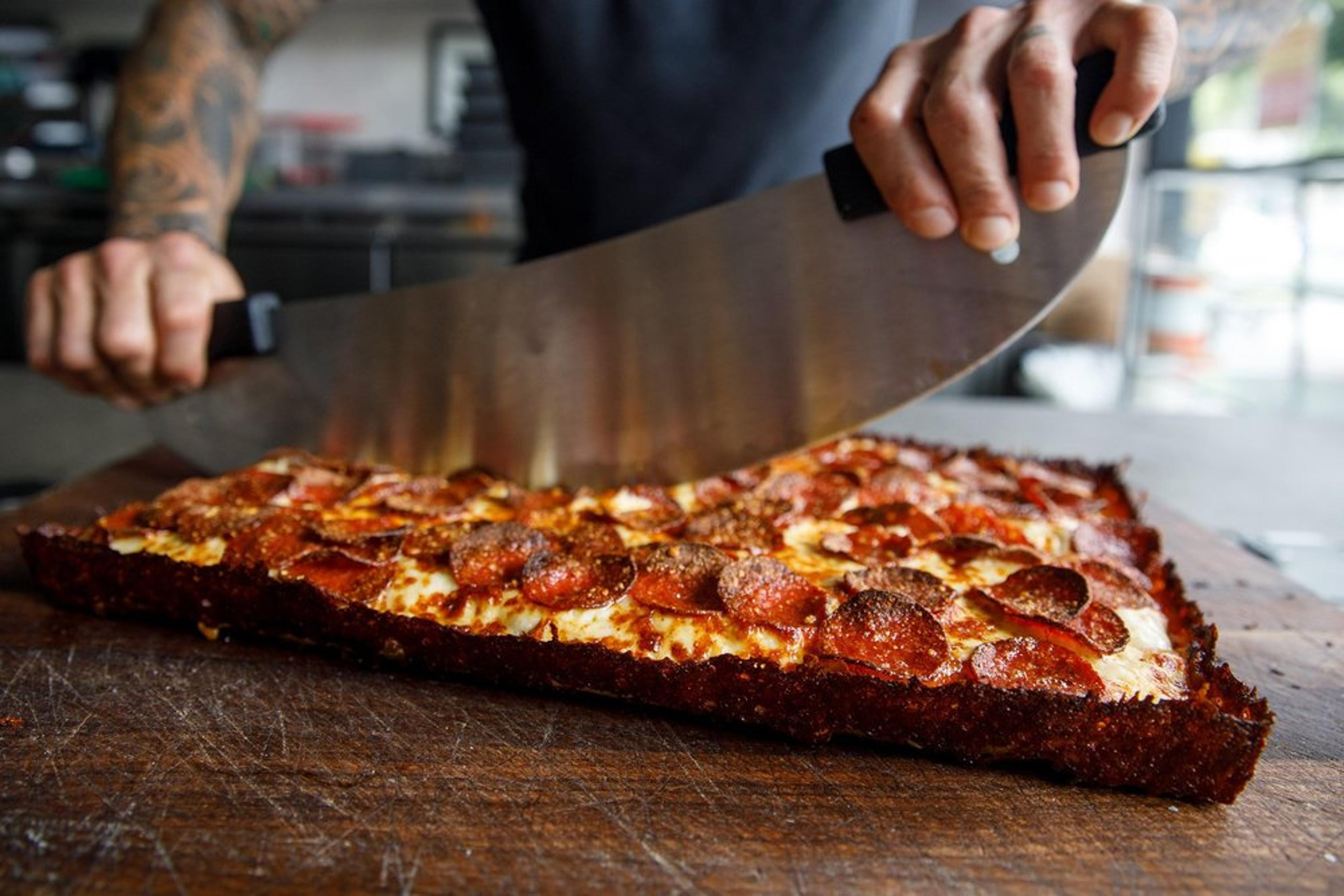 A Detroit-style pepperoni pizza being sliced with a big rocking knife.