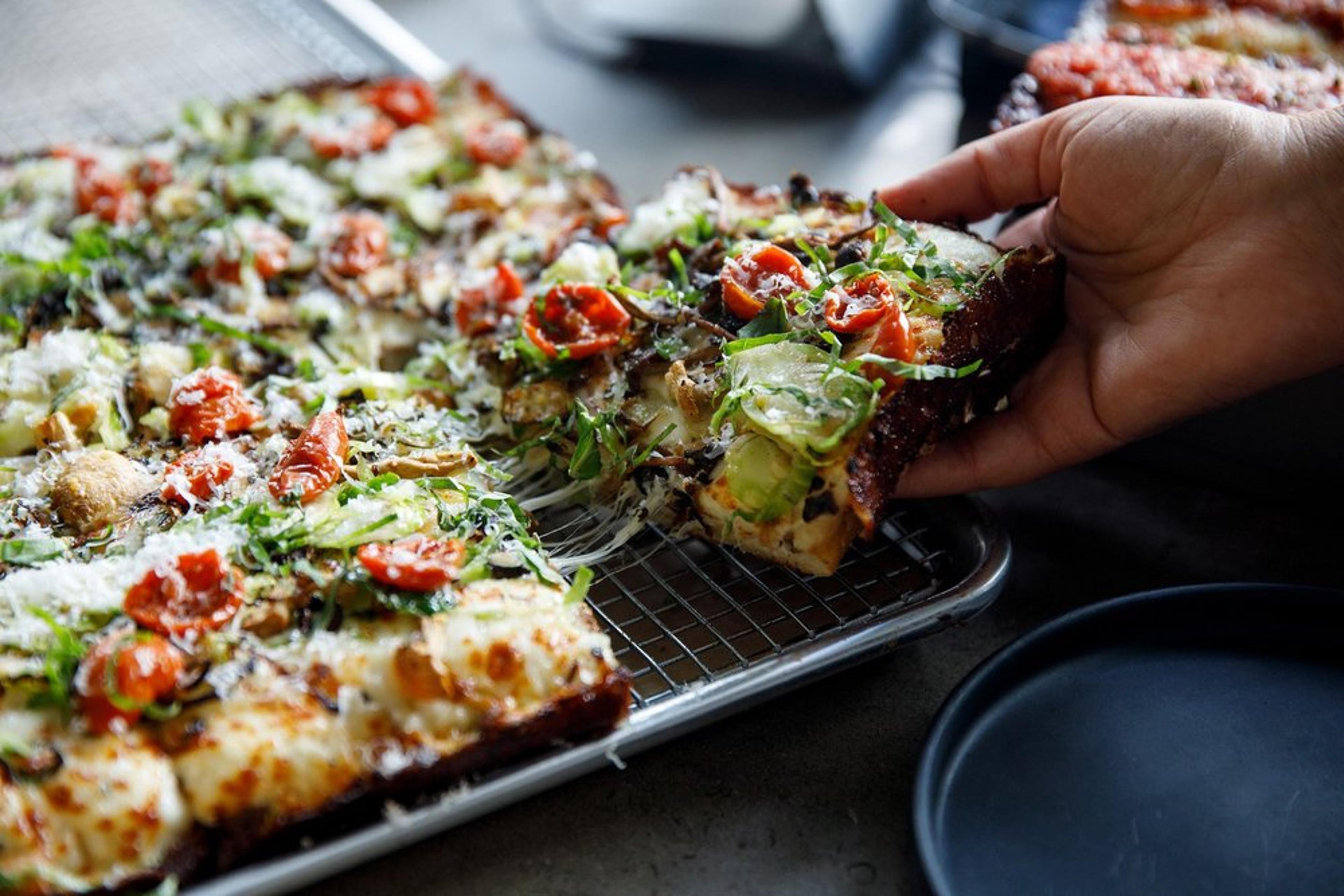 A person lifts a slice of rectangular pizza from a baking rack. The pizza is topped with cheese, sliced tomatoes, and greens. A black plate is partially visible in the foreground.