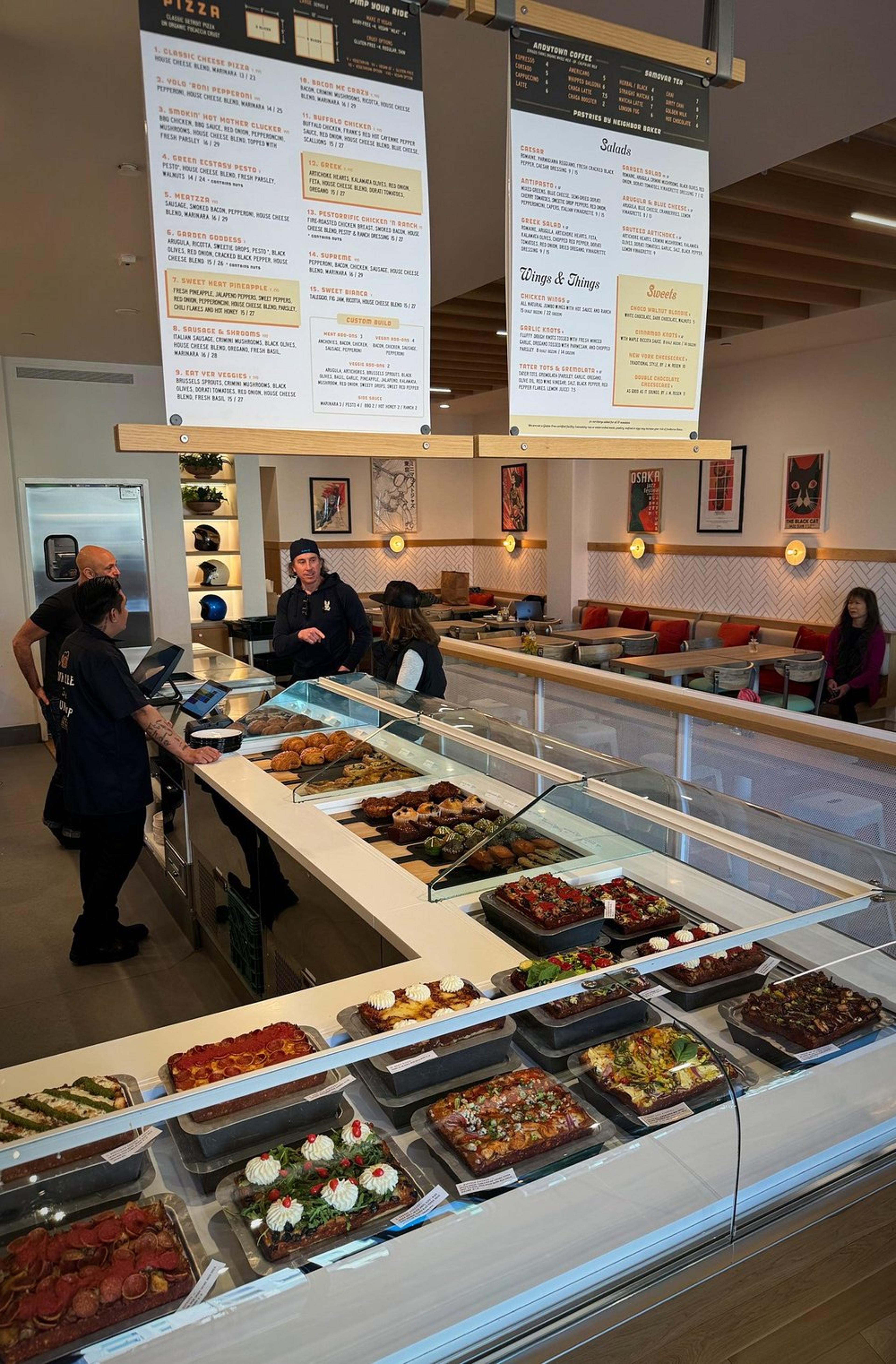 Joyride Pizza display case with a variety of Detroit-style pizzas inside a modern cafe or bakery. The background shows a counter area with staff and customers, and large menu boards hanging from the ceiling.