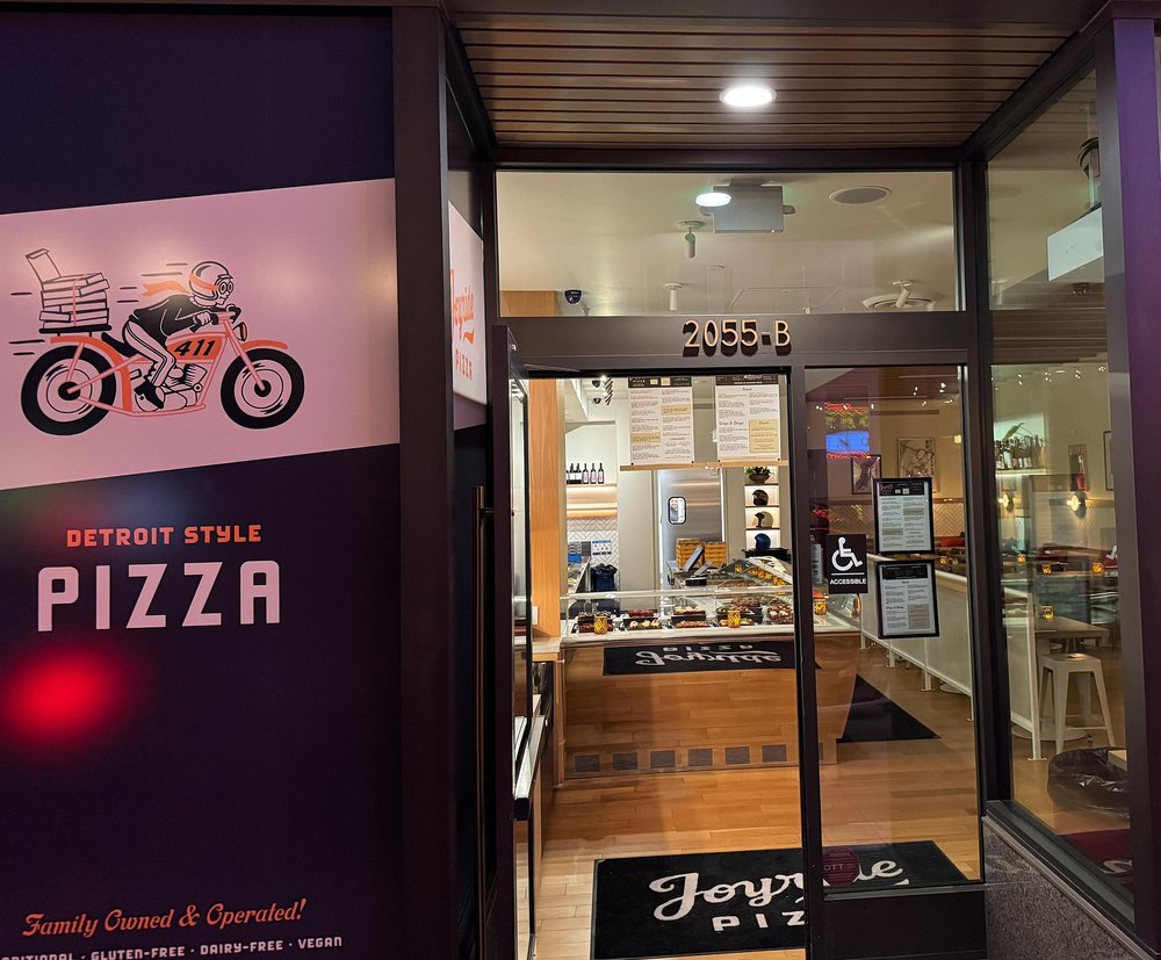 Entrance of Joyride Pizza in the Marina with a sign showing a motorcyclist with a delivery pizza box, located at 2055-B, with interior visible including menu boards, display case with food, and a seating area.