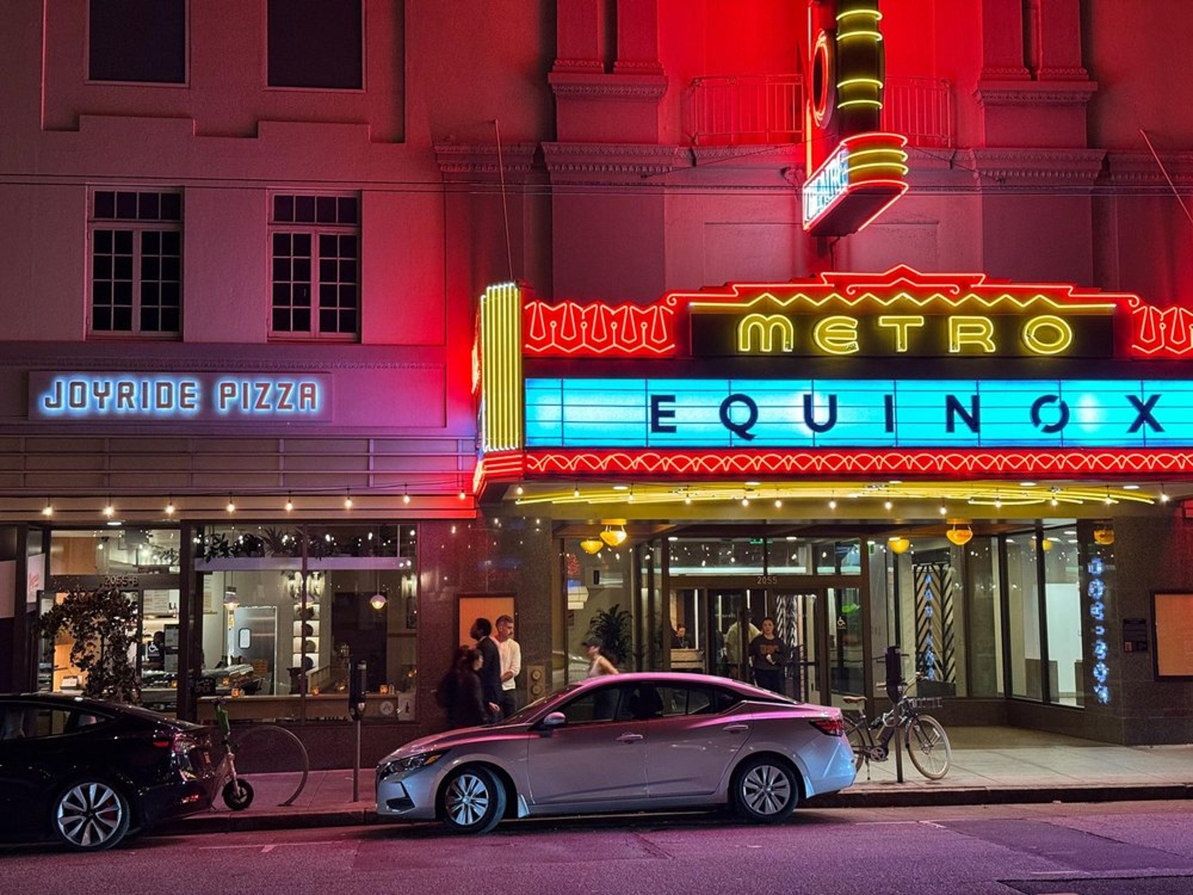 A street scene at night shows the illuminated facade of the Metro theater with the movie title Equinox. Next to it is Joyride Pizza with a brightly lit sign. Several cars are parked on the road, and a bicycle is in front of the theater.