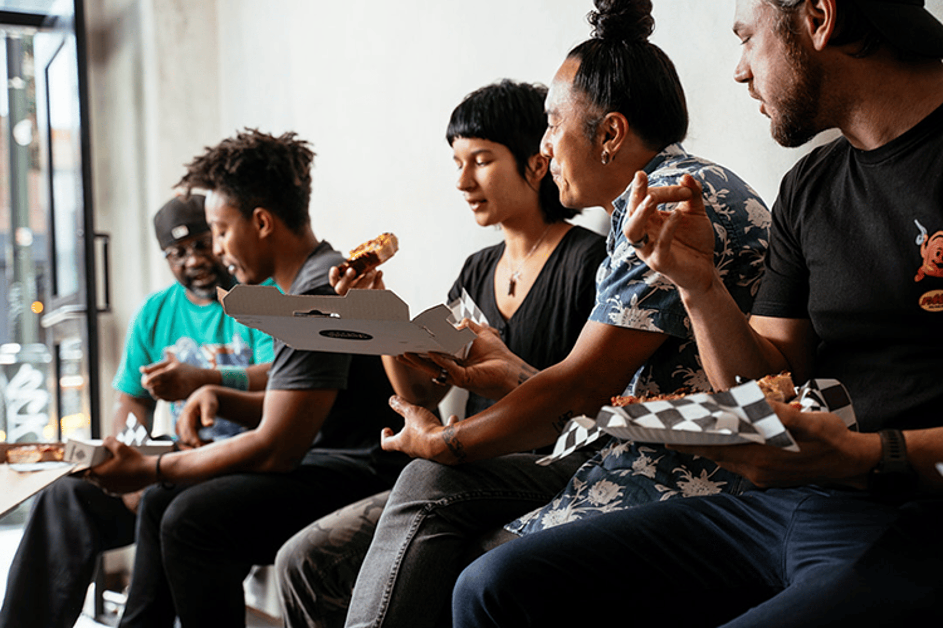 Friends sitting side by side by the window, eating slices of Joyride pizza from paper boats.