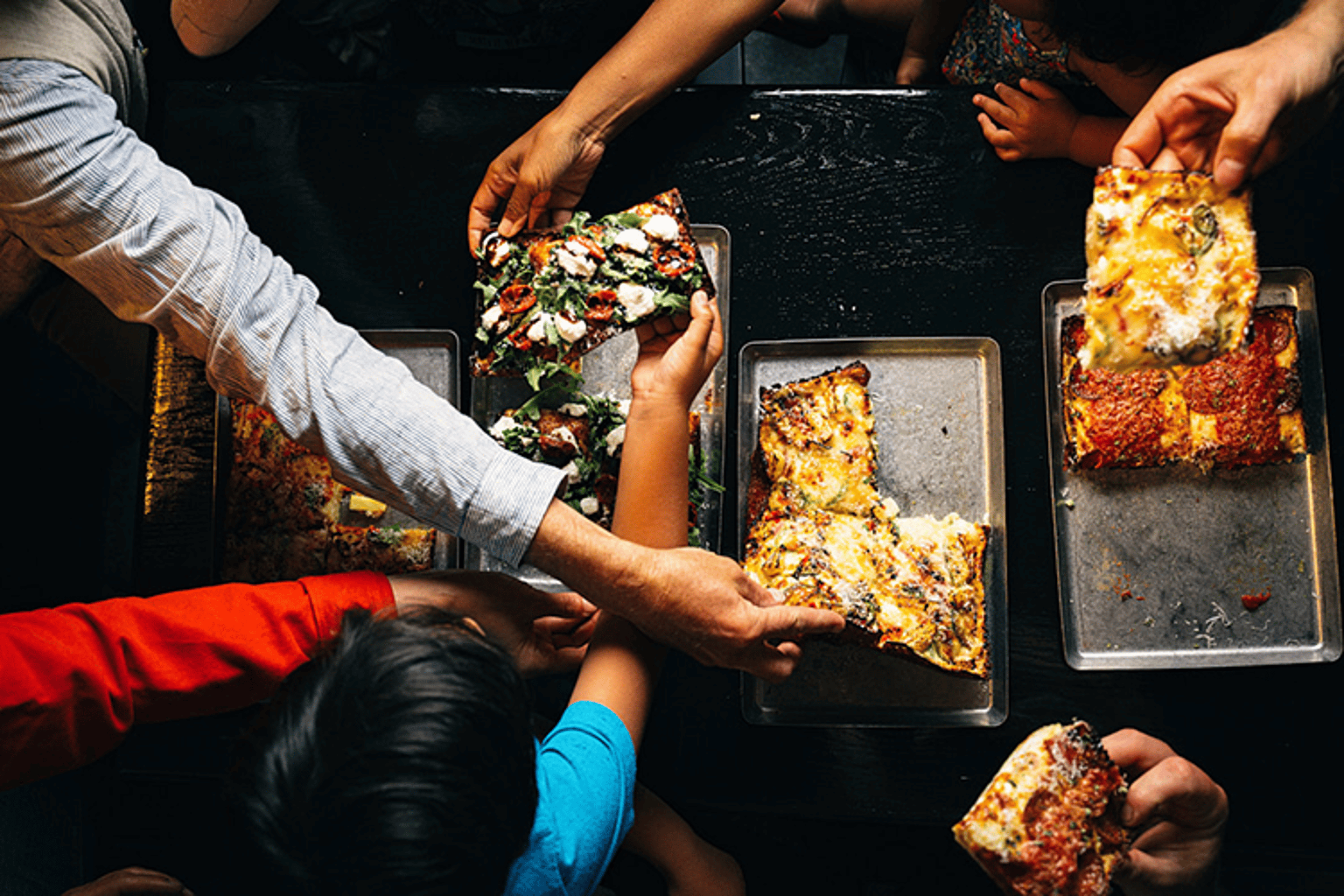 Overhead view of hands reaching for slices from trays of Detroit-style pizza at a party.