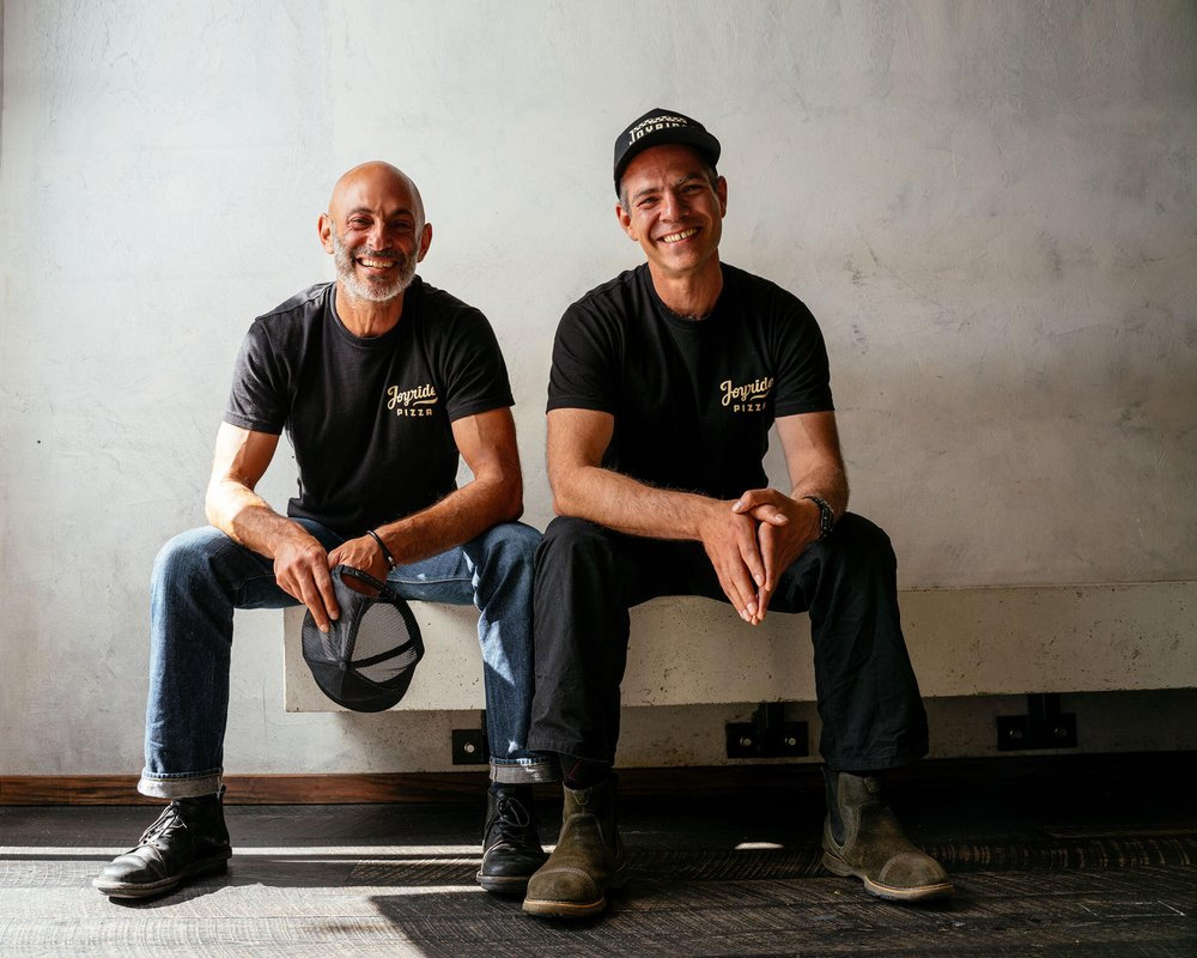 Two smiling men in matching black Joyride t-shirts sit on a bench against a tiled wall.