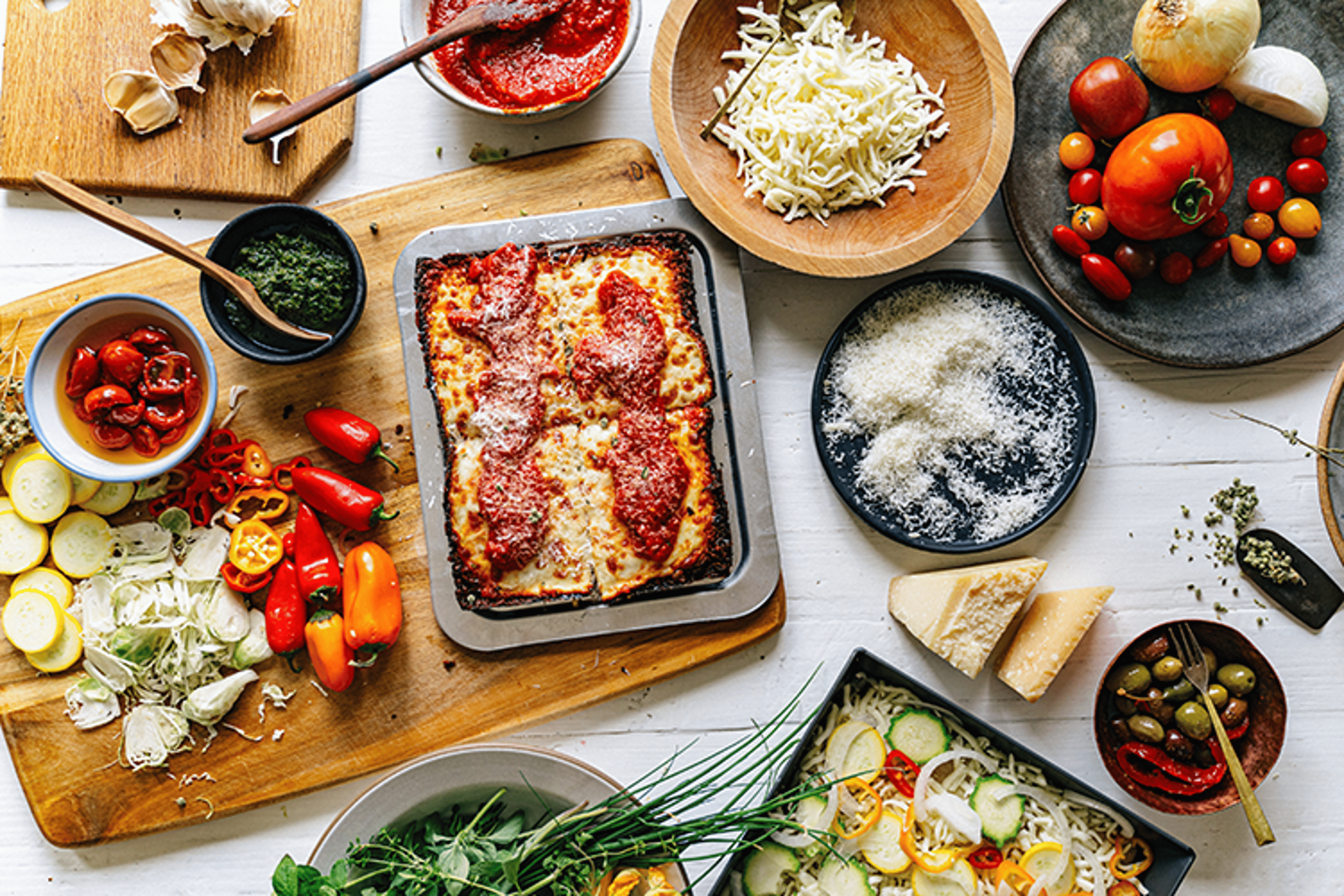 Fresh tomatoes, peppers, herbs, cheeses and olives laid out around a pan of Detroit-style pizza.