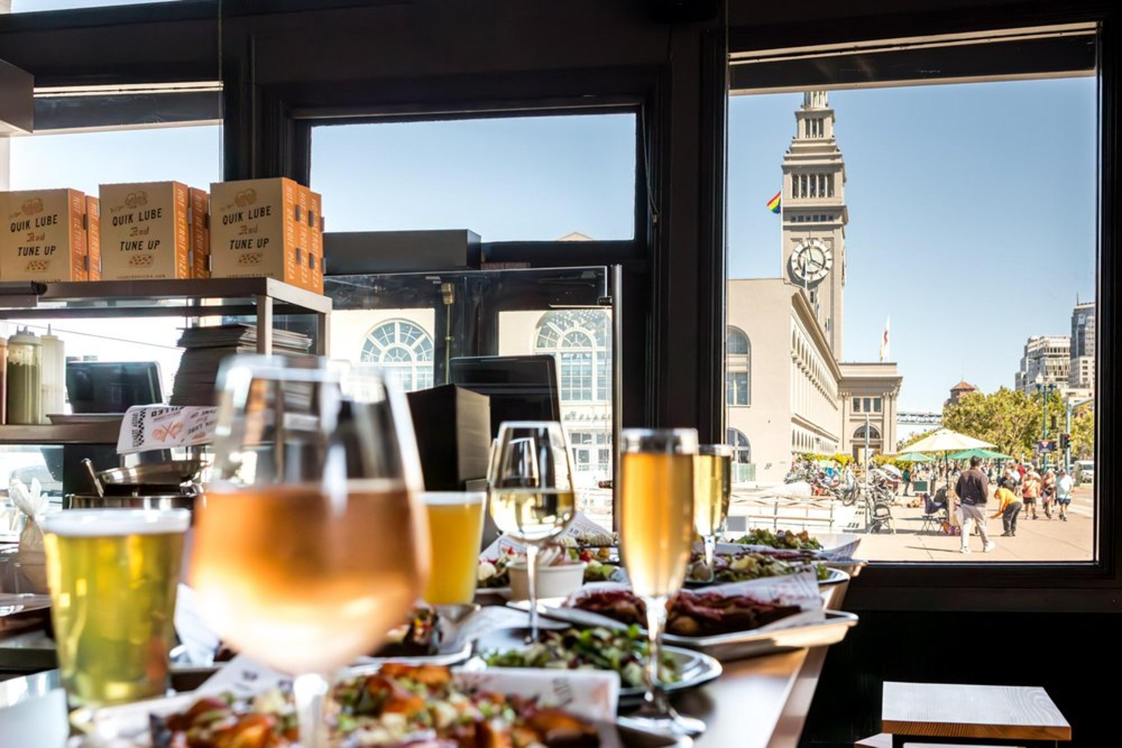 A table with glasses of wine and plates of food overlooks a window view of a city scene, featuring a clock tower and bustling plaza outside.