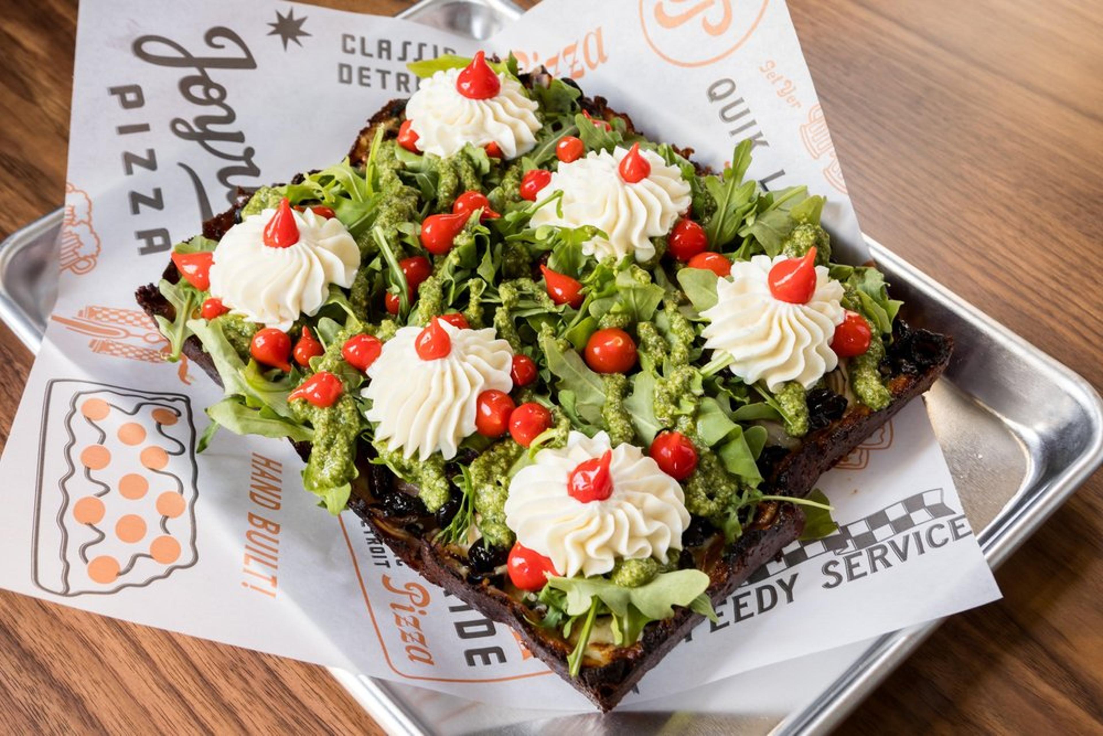 Detroit-style square pizza topped with arugula, sweety-drops, dollops of white whipped topping with red sauce accents, green pesto drizzle, and a toasted crust, served on a metal tray with branded paper