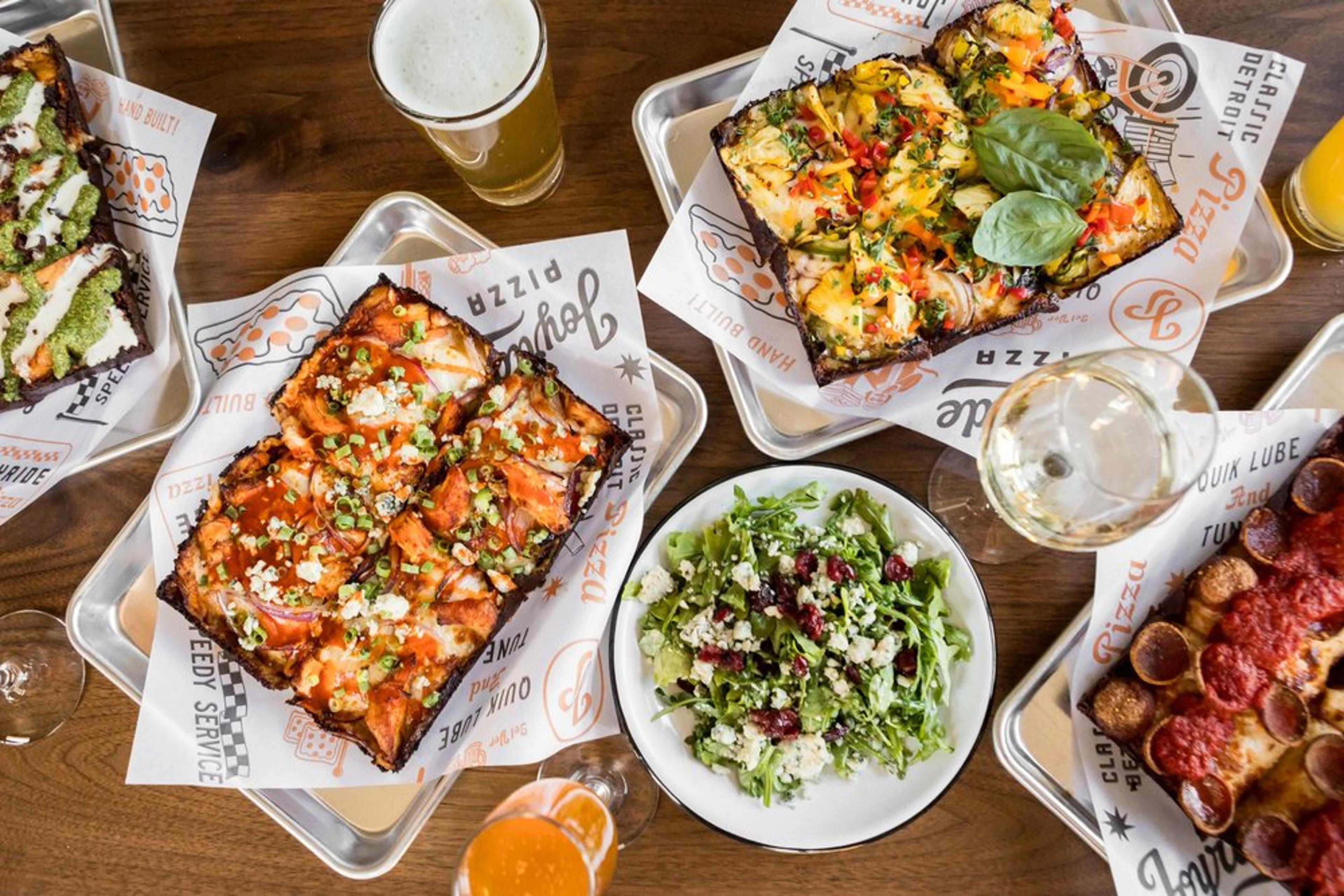Top-down view of a table with four Detroit-style pizzas on trays, a salad, and several drinks including beers, wine, and orange juice.