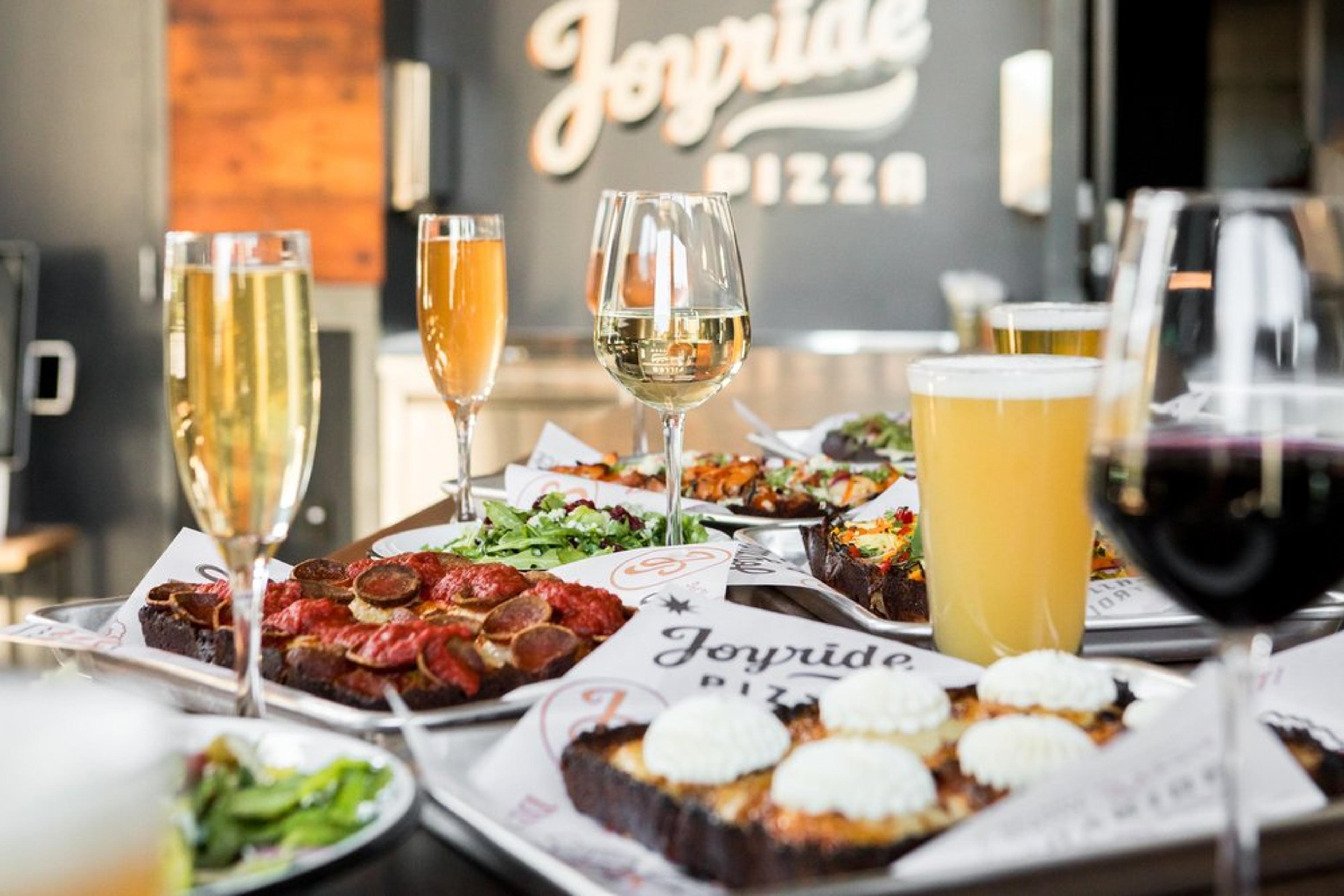 A table at Joyride Pizza on Pier 1 with Detroit-style pizzas, salads, and drinks at a restaurant, with a sign for Joyride Pizza in the background.