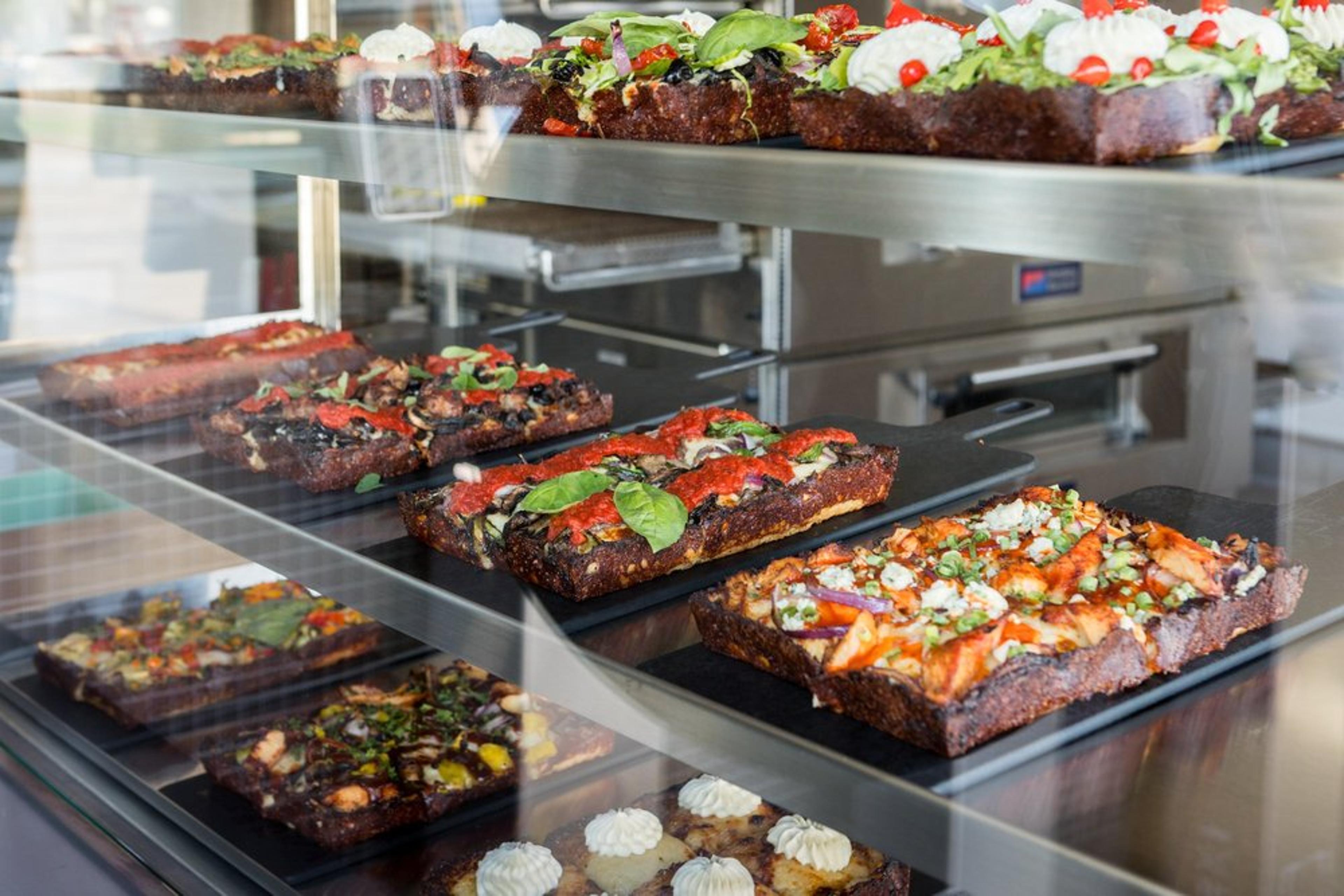 Variety of rectangular pizza slices with different toppings displayed in a glass case, including vegetables and cheese.