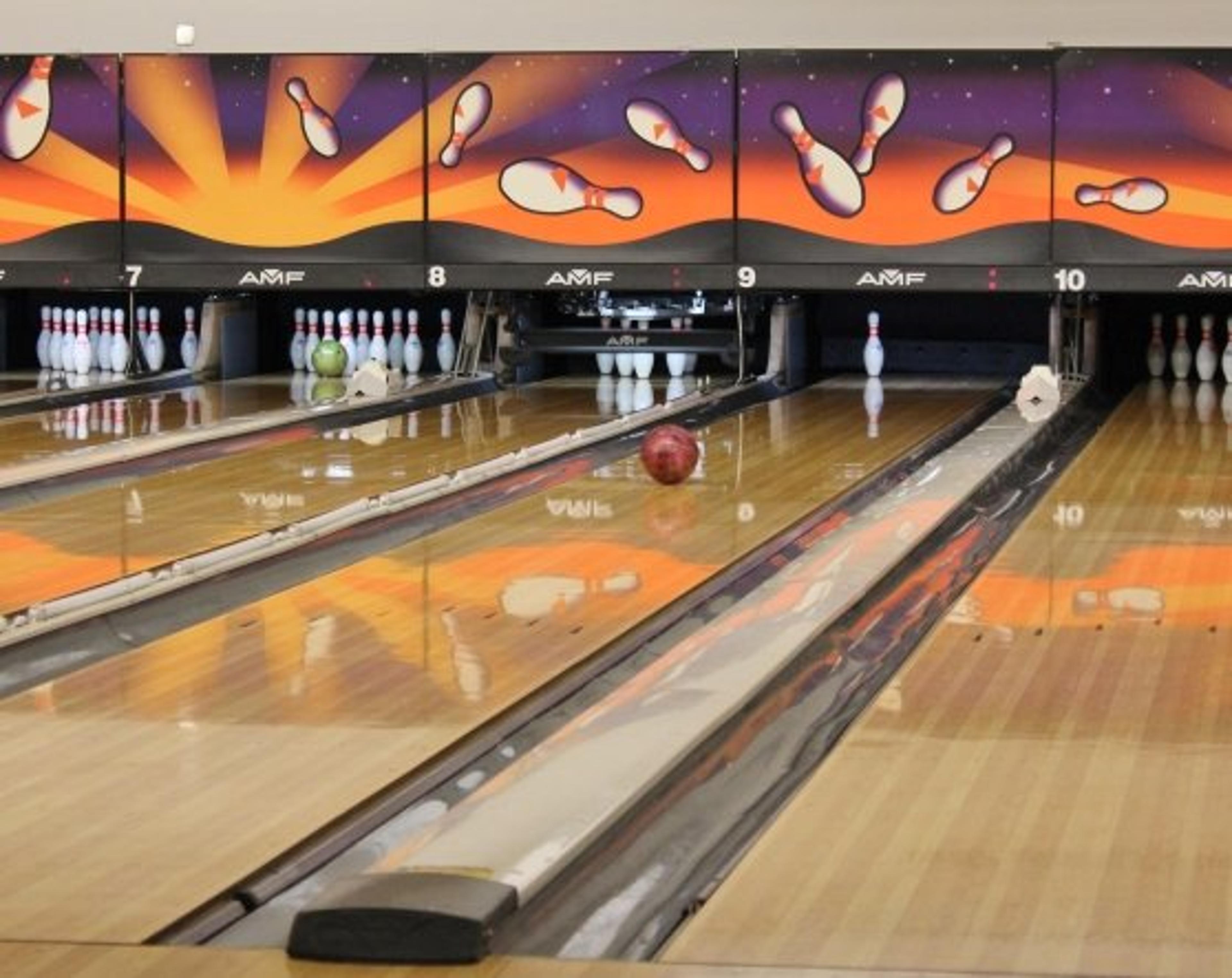 A bowling alley with three lanes. A green ball is rolling toward pins on the left lane, a red ball on the middle lane, and another red ball on the right lane. The lanes are wooden with decorative panels above the pins.