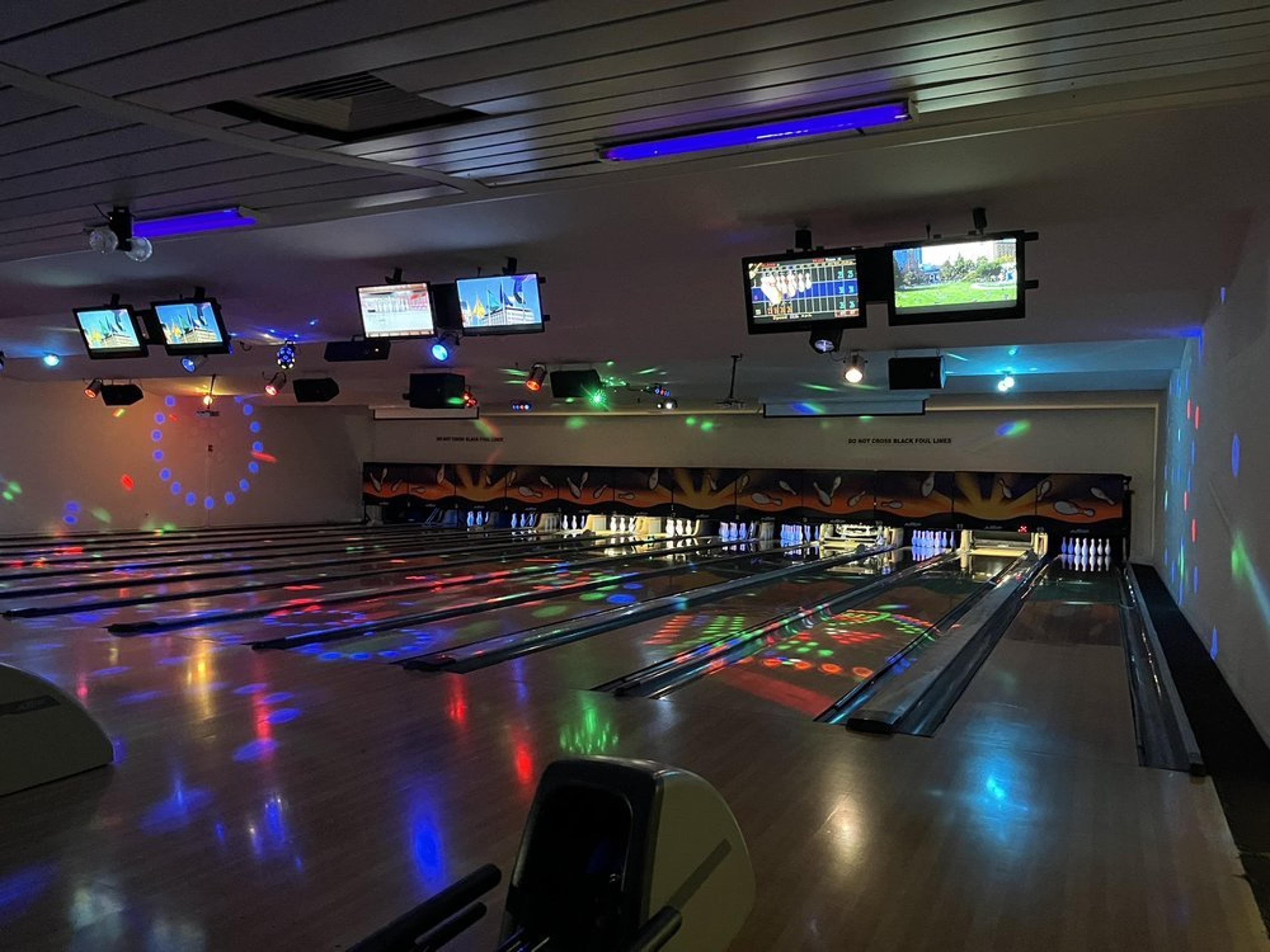 A dimly lit bowling alley with several lanes and colorful lights reflecting on the floor. Monitors above display games, and a clock with numbered pins is seen on the left wall.