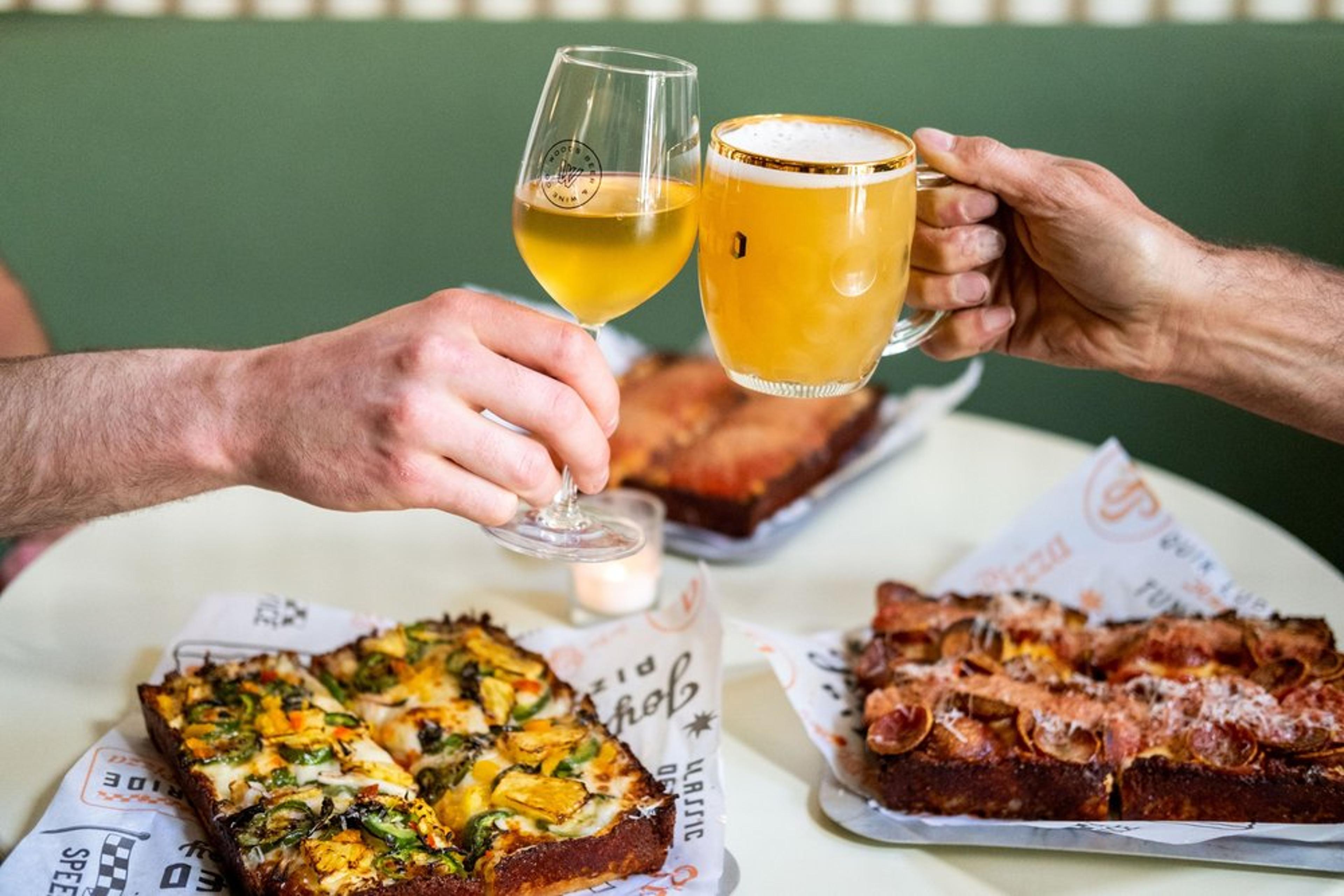 Two friends clink glasses of beer over trays of Detroit-style pizza