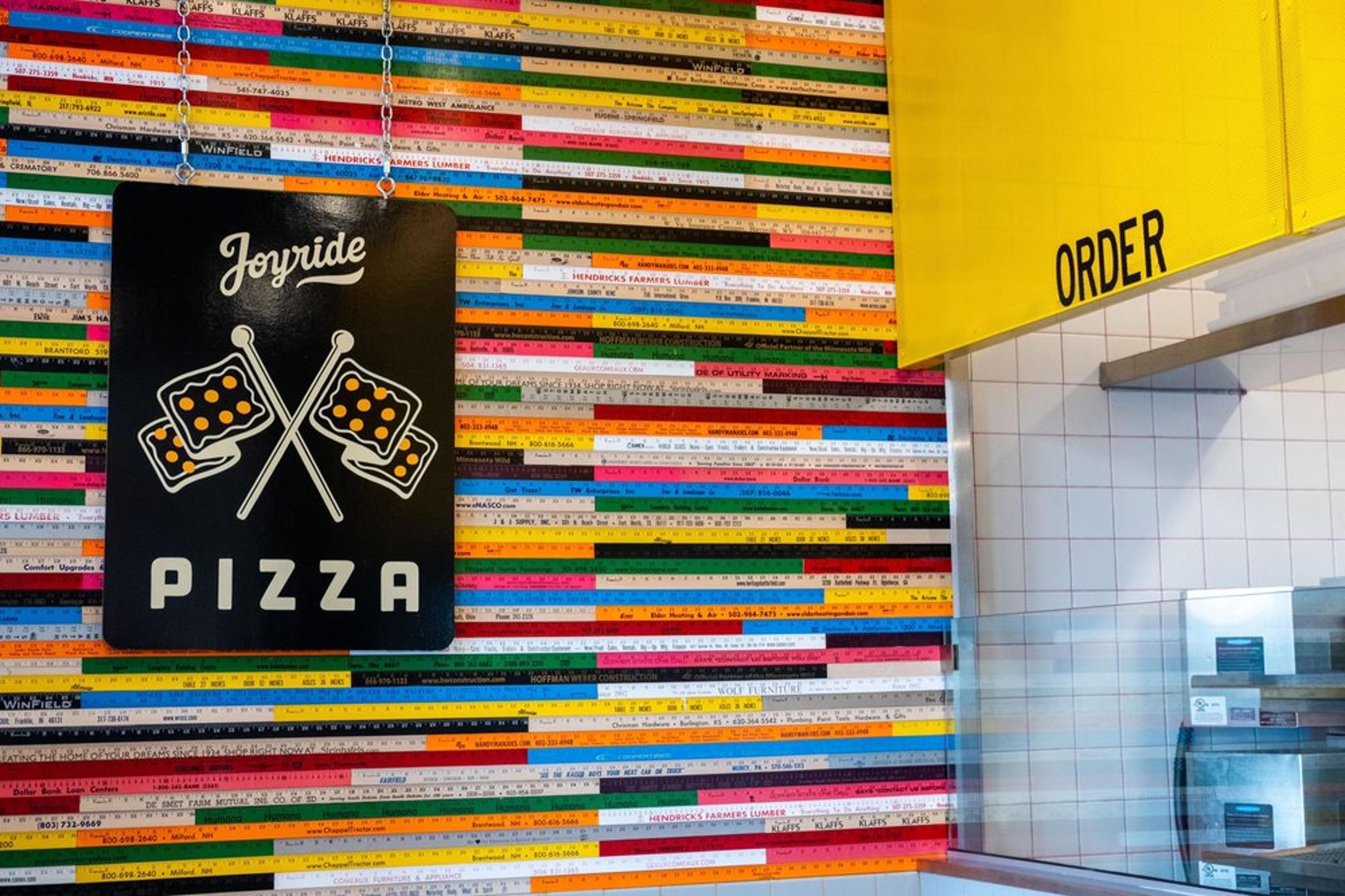 Colorful wall with wooden rulers and a black sign for Joyride Pizza featuring crossed pizza flags. A yellow menu board on the right labeled ORDER above a counter.