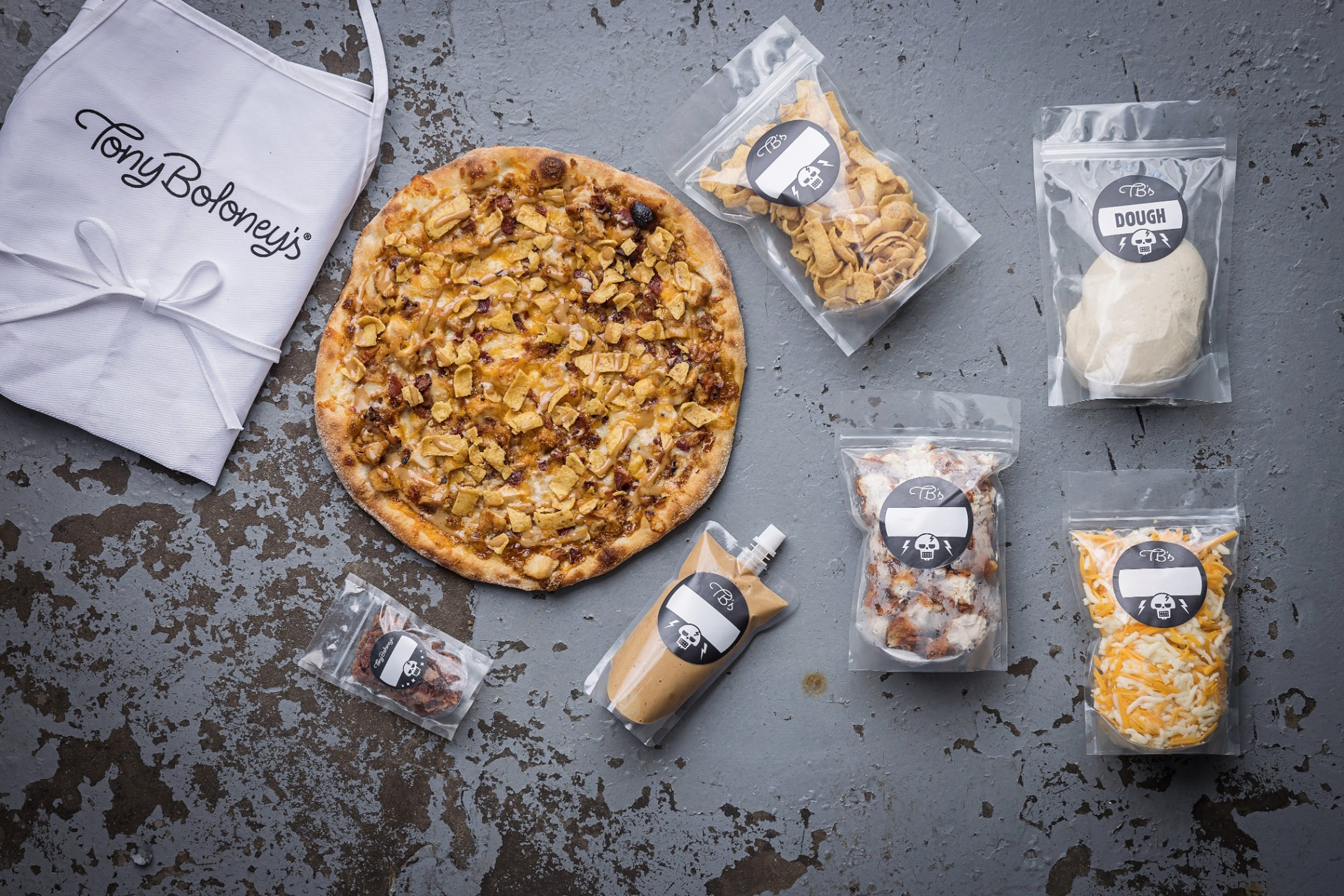 A Tony Boloney's apron, a finished pizza, and labeled kit ingredients laid out