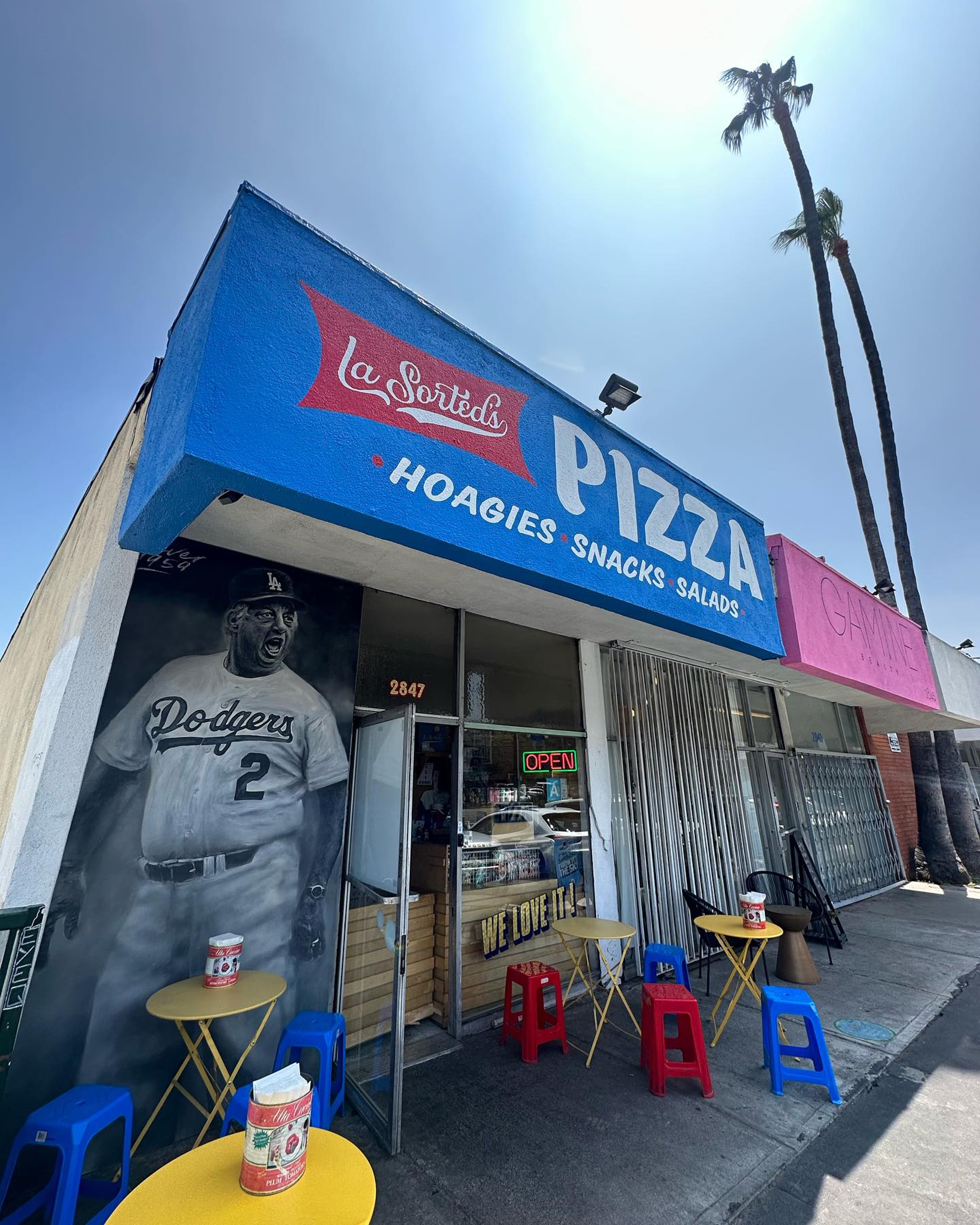 La Sorted's Pizza storefront with outdoor yellow tables, red and blue stools, and a mural of a Dodgers player wearing jersey number 2.