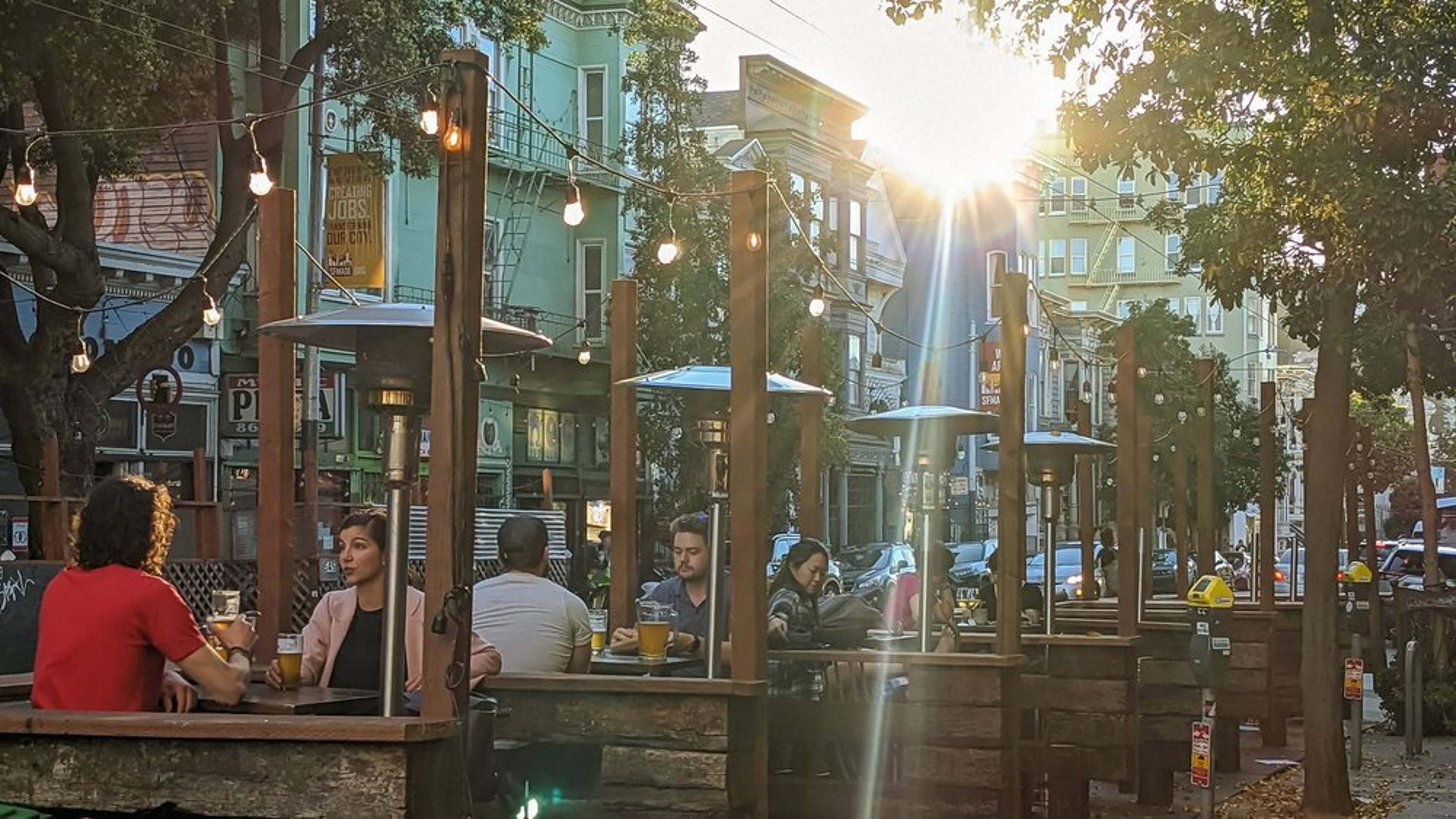 People sit and enjoy drinks at outdoor wooden tables on a street patio during sunset. String lights hang above, and trees line the sidewalk. Sunlight filters through, creating a warm ambiance in an urban setting.