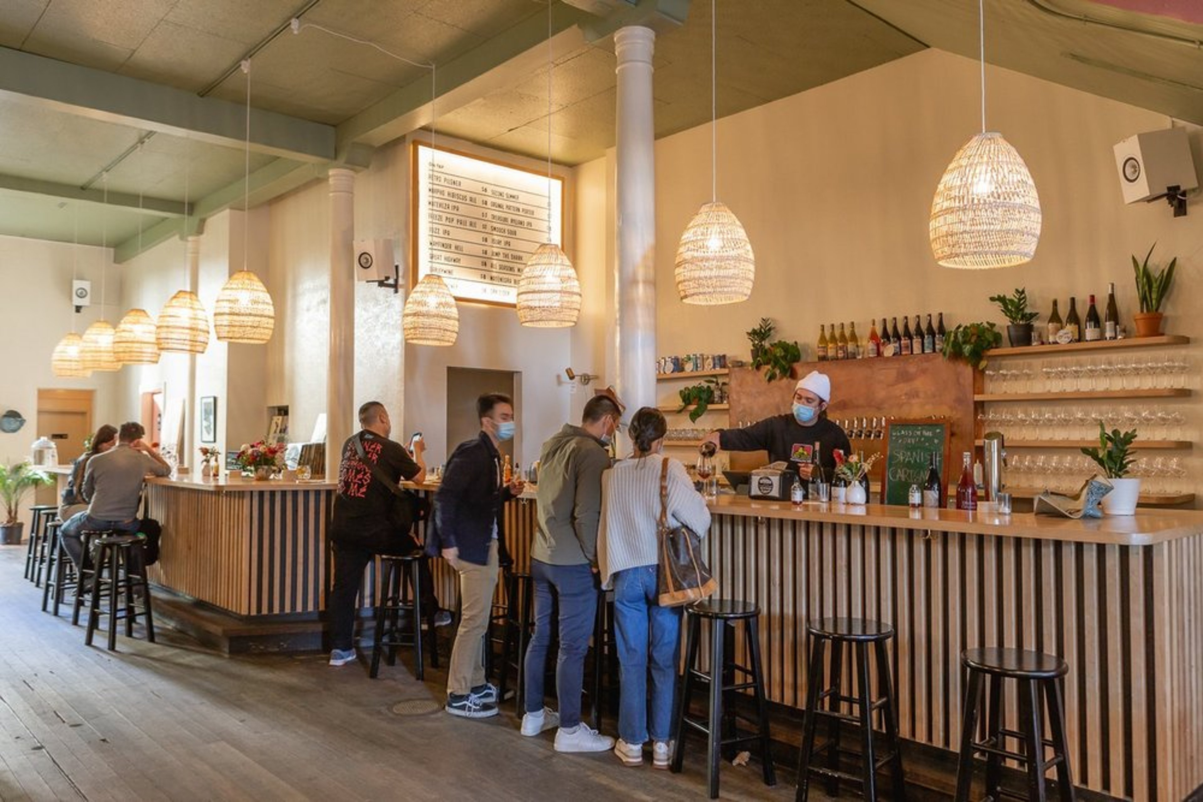 People are lined up at a stylish bar with pendant lights and wooden decor. A bartender serves drinks while patrons, some wearing masks, chat and wait. The bars menu is displayed overhead. Wall shelves hold bottles and small plants.