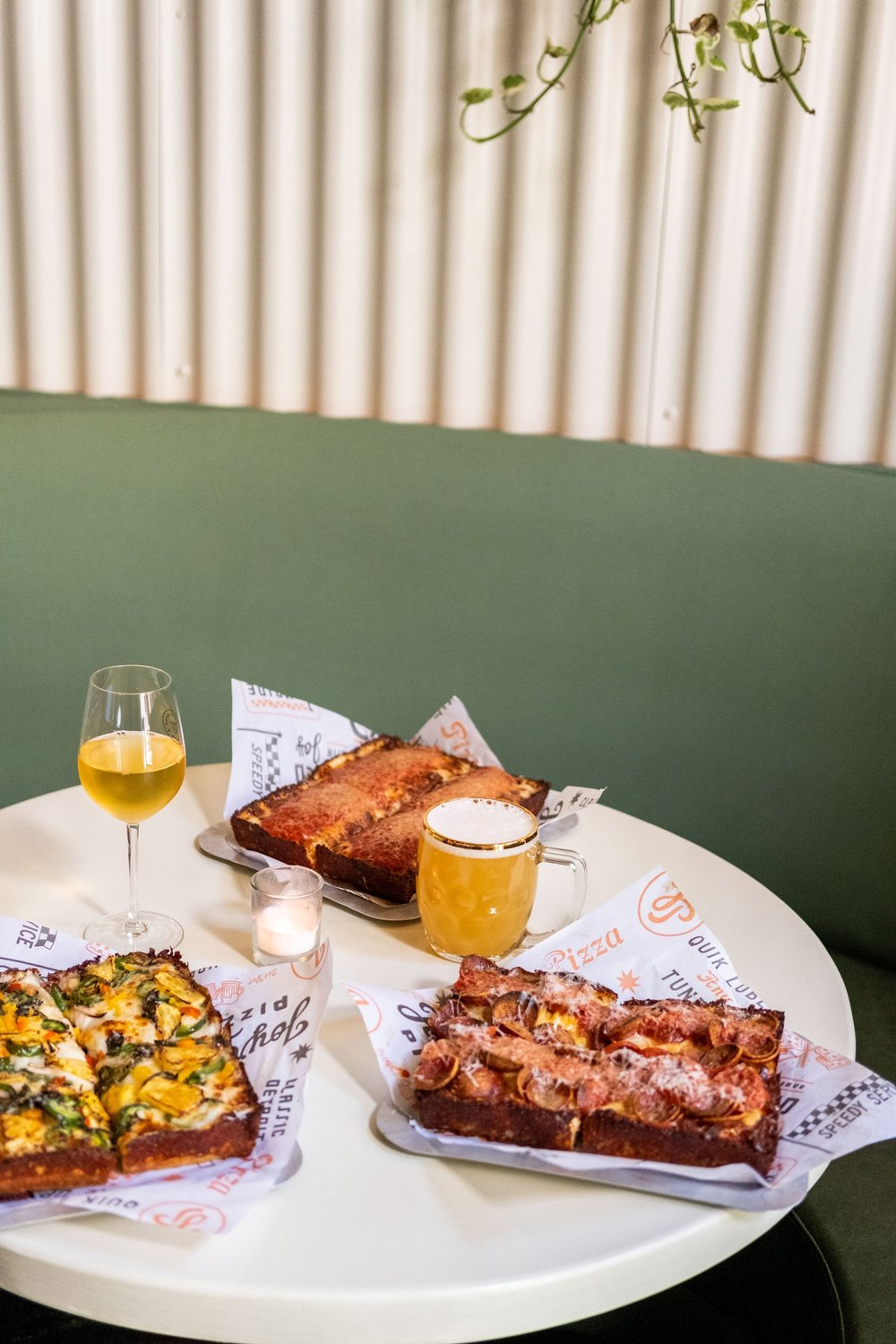 A table with three pizzas served on trays, a beer mug, and a glass of white wine. The pizzas have various toppings. The setting is cozy with a green cushioned seat in the background. A lit candle adds warmth.
