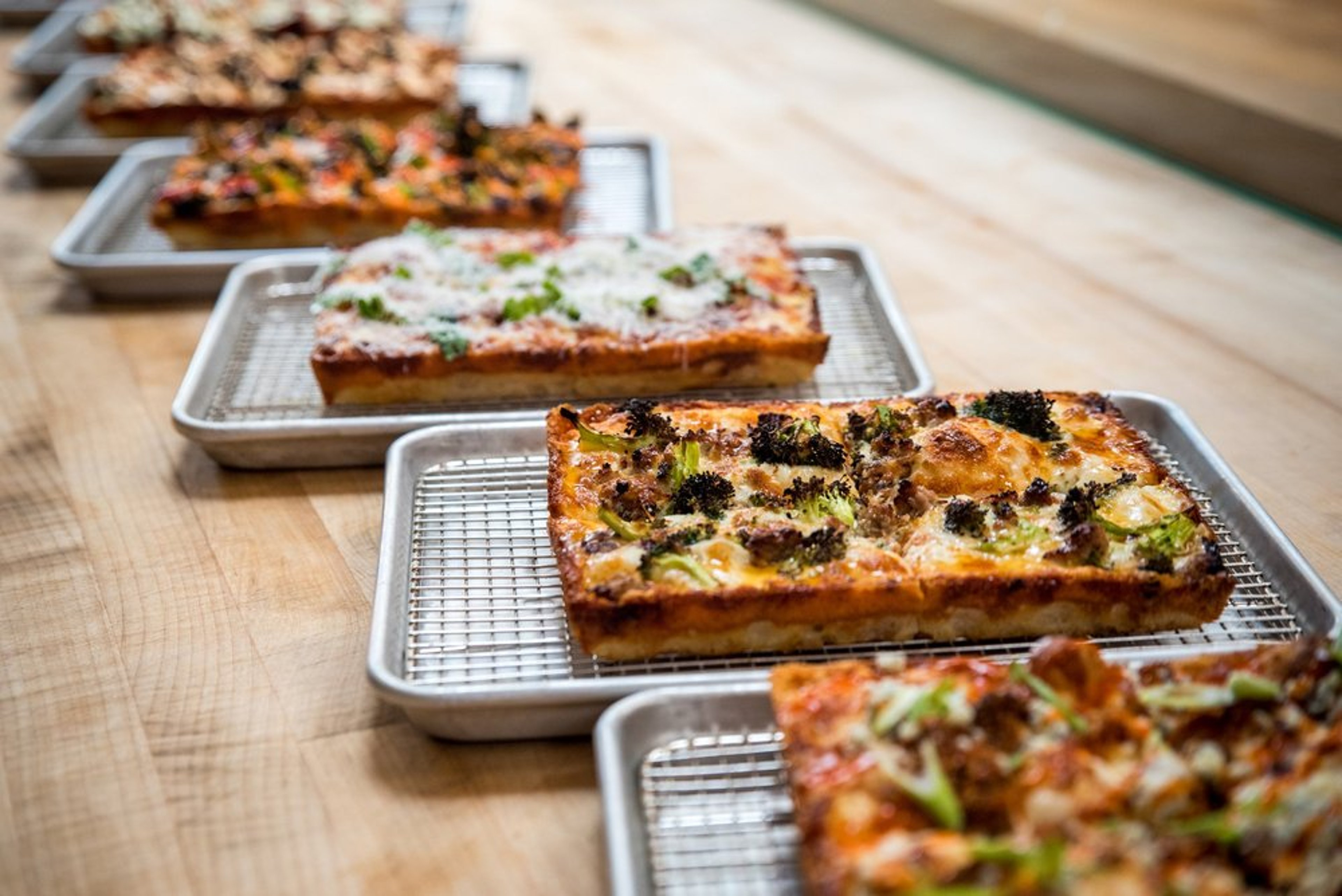 Four trays of different varieties of rectangular pizza on a wooden table.