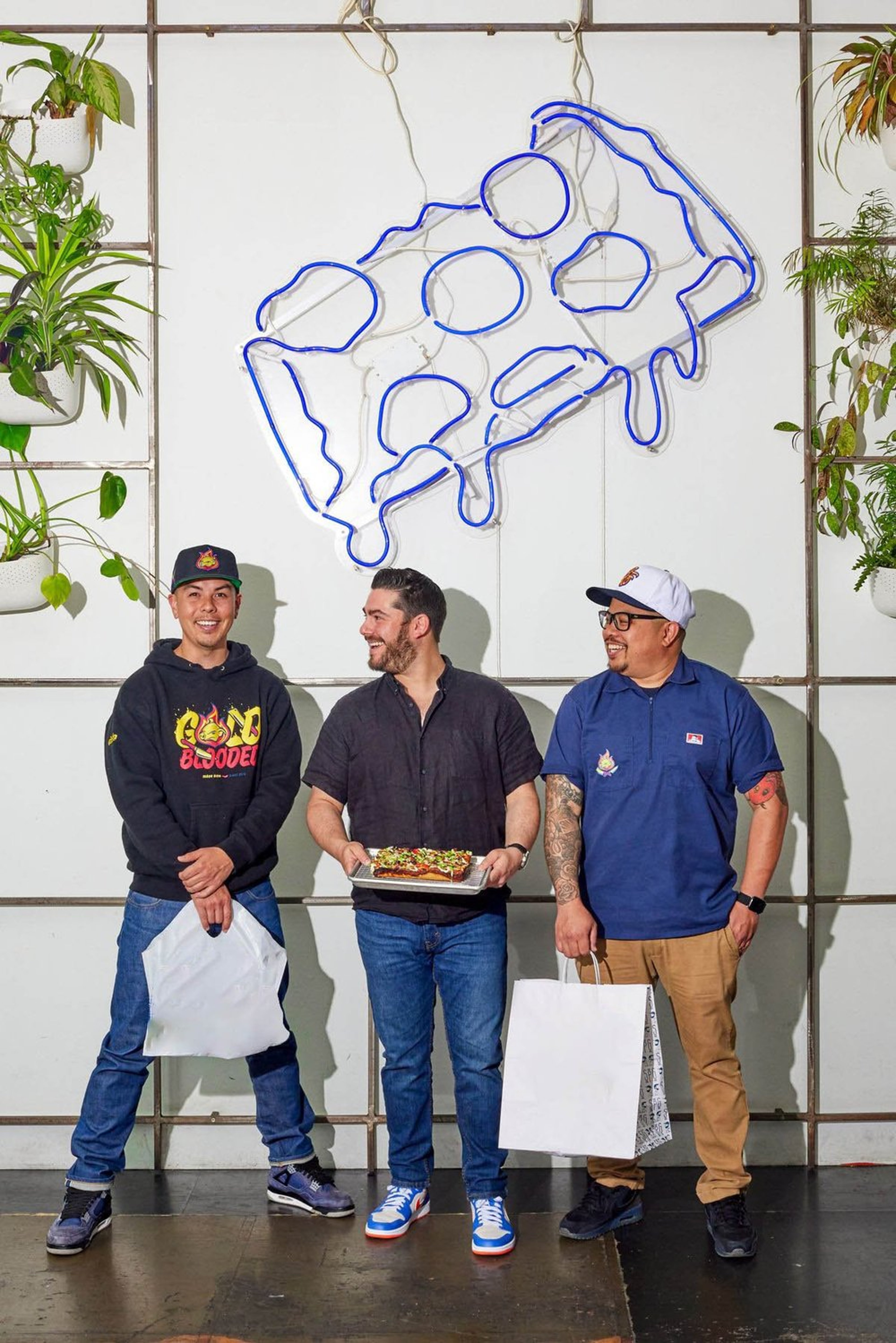 Three men standing indoors in front of a white wall with a blue neon line art of a landmass. The man in the middle holds a tray with a decorated pizza, while the other two carry shopping bags. All are smiling and looking at each other.
