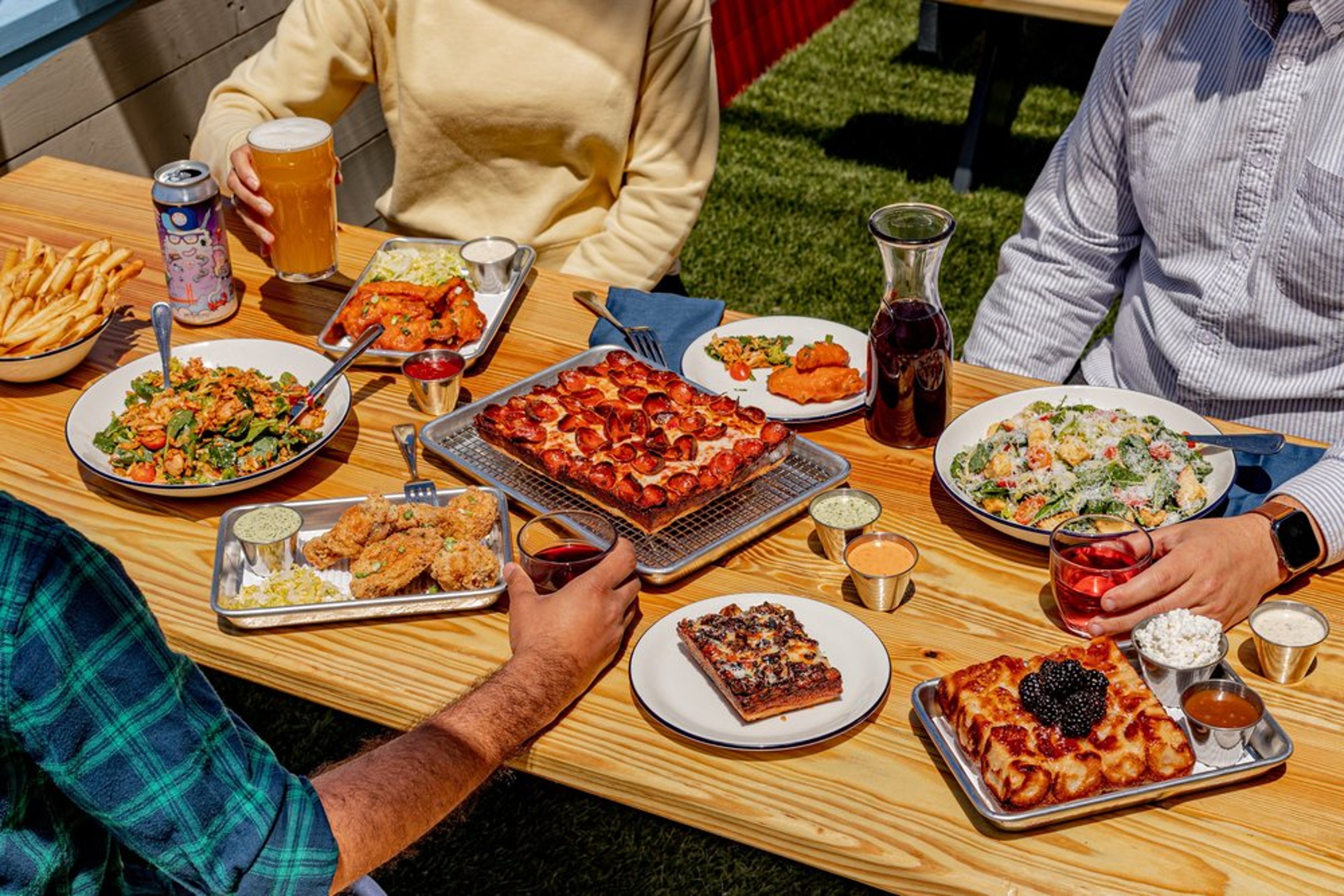 People enjoying a meal outdoors with various dishes including pizza, salad, fried chicken, and drinks on a wooden table.