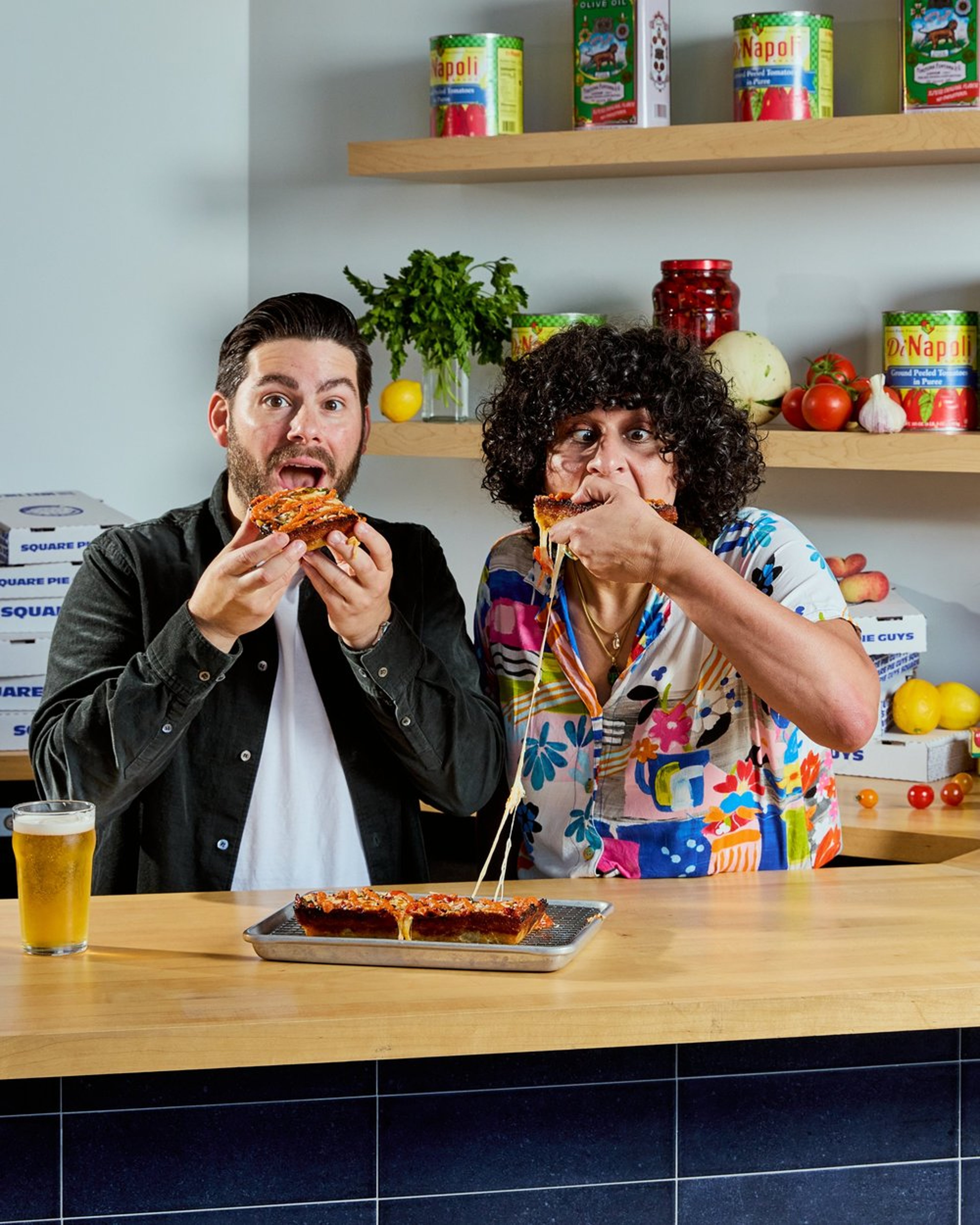 Two people eating pizza in a kitchen with shelves of canned goods, fresh vegetables, and a lemon plant in the background, with a glass of beer on the table.
