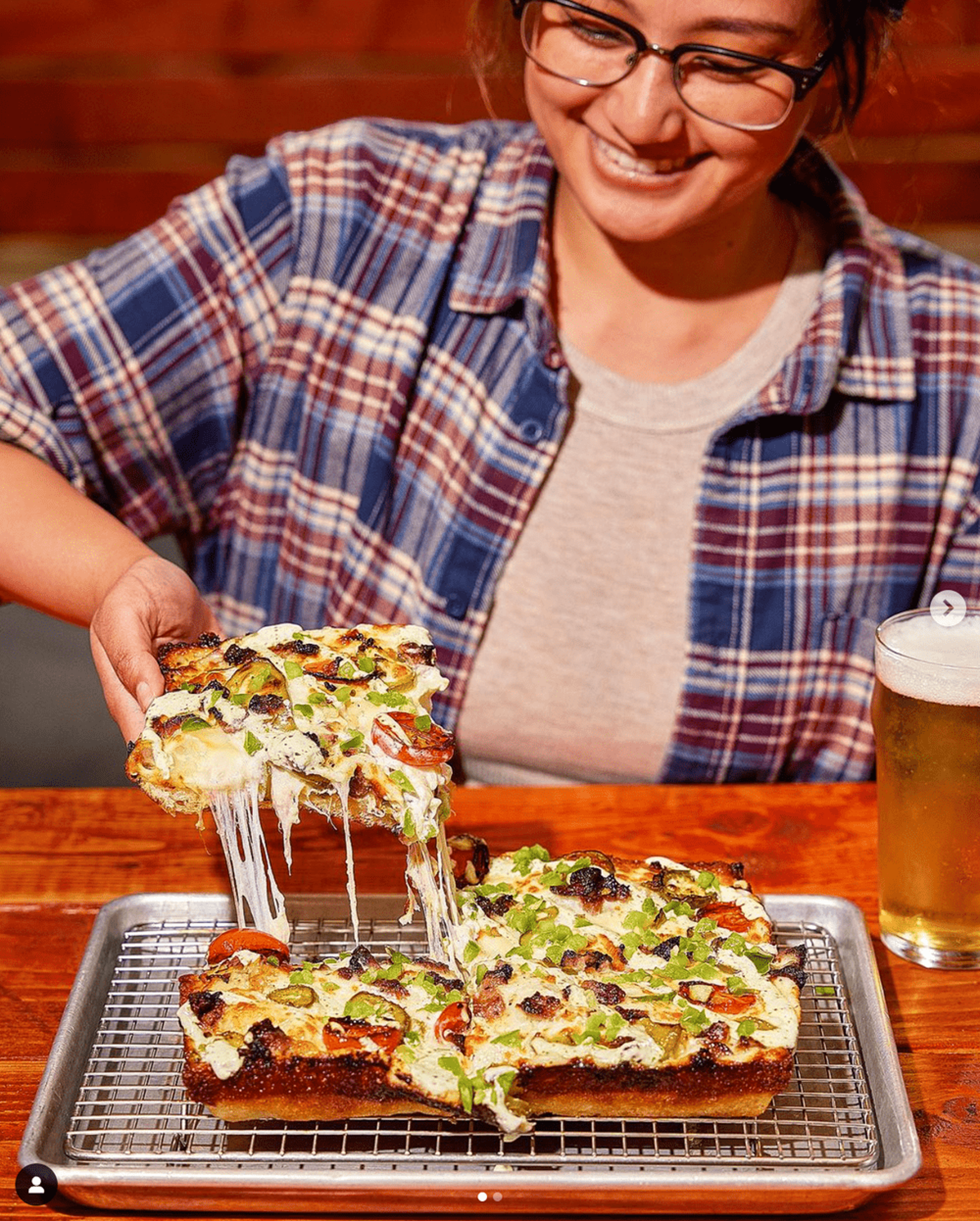 A woman with glasses and a plaid shirt smiling as she pulls a slice of pizza topped with vegetables from a tray, on a wooden table with a glass of beer nearby.