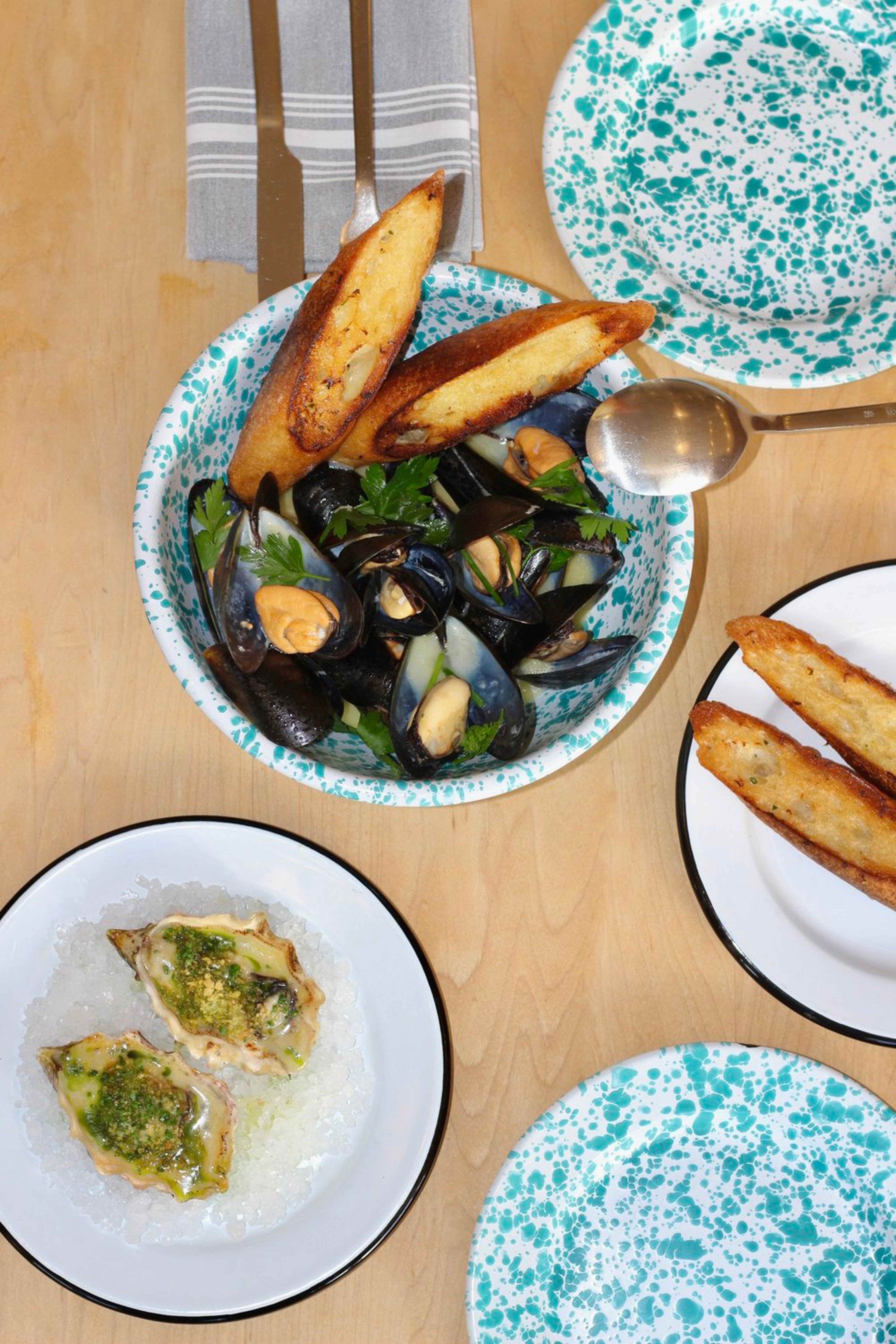 A table with a dish of cooked mussels garnished with green herbs, two toasted bread slices, and a plate of oysters topped with green sauce.