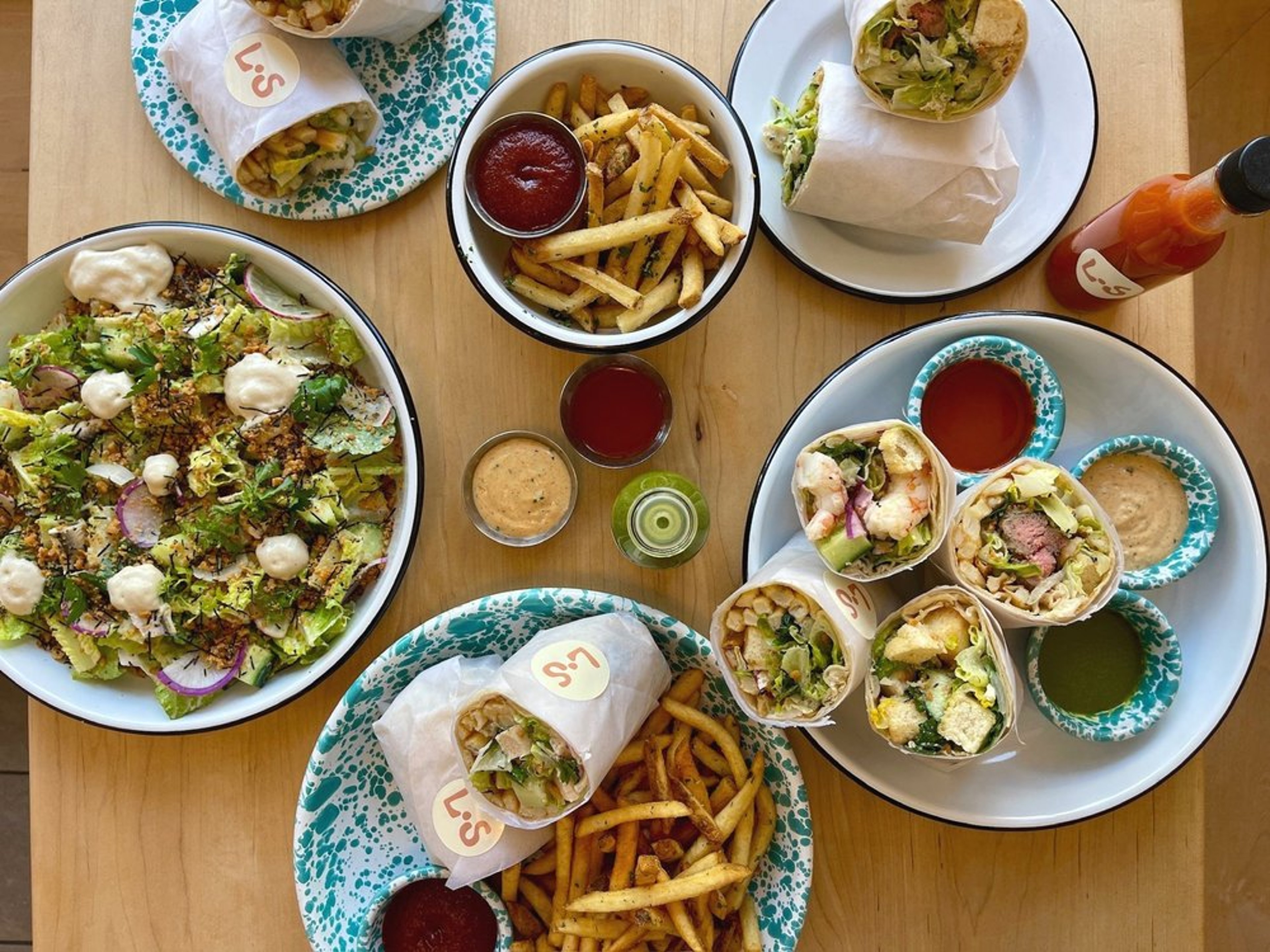 Overhead view of the Little Shucker counter with caesar salads, wraps, fries, and sauces on speckled plates.