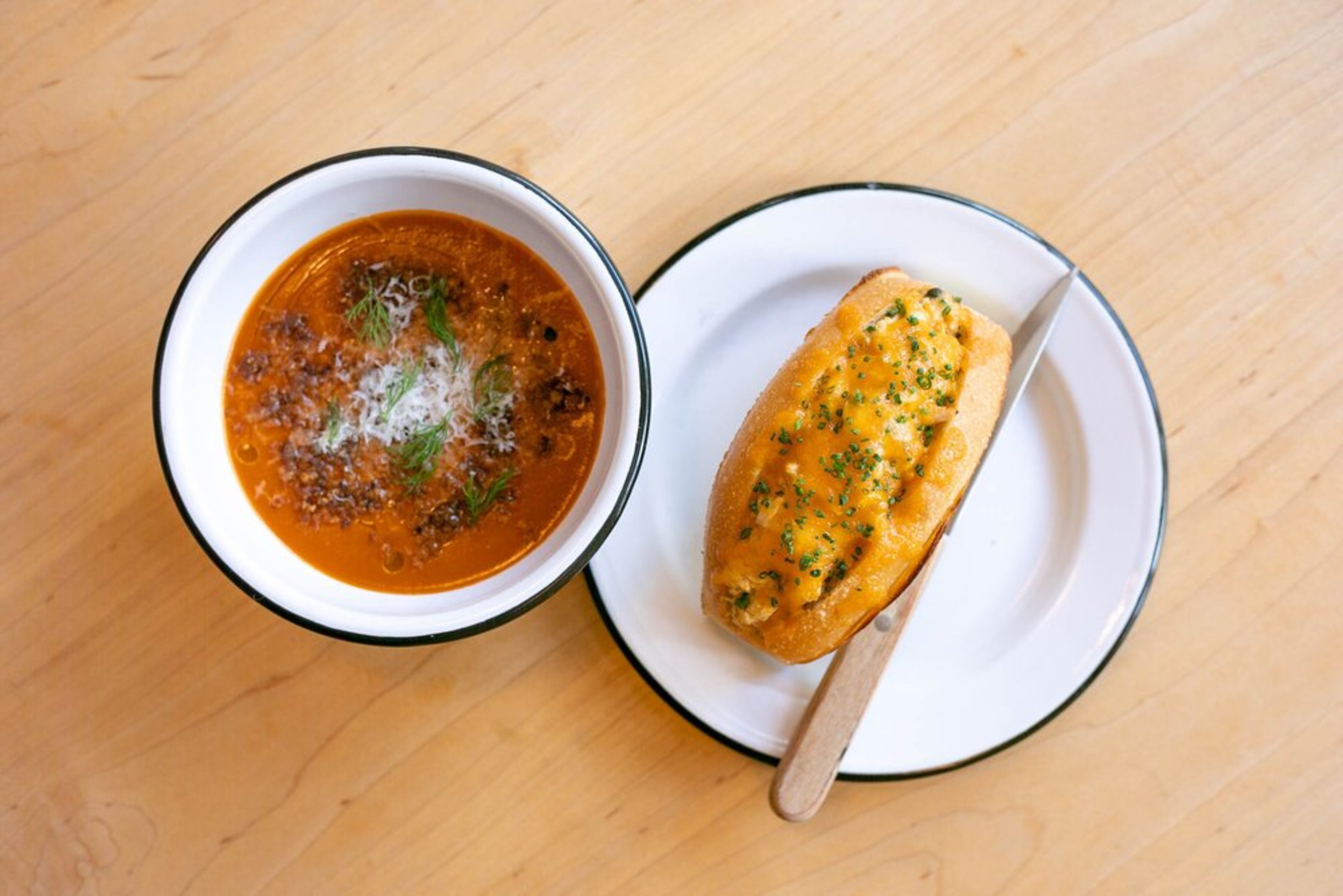 A bowl of tomato soup garnished with herbs and cheese, next to a plate with a piece of garlic bread topped with melted cheese and herbs.
