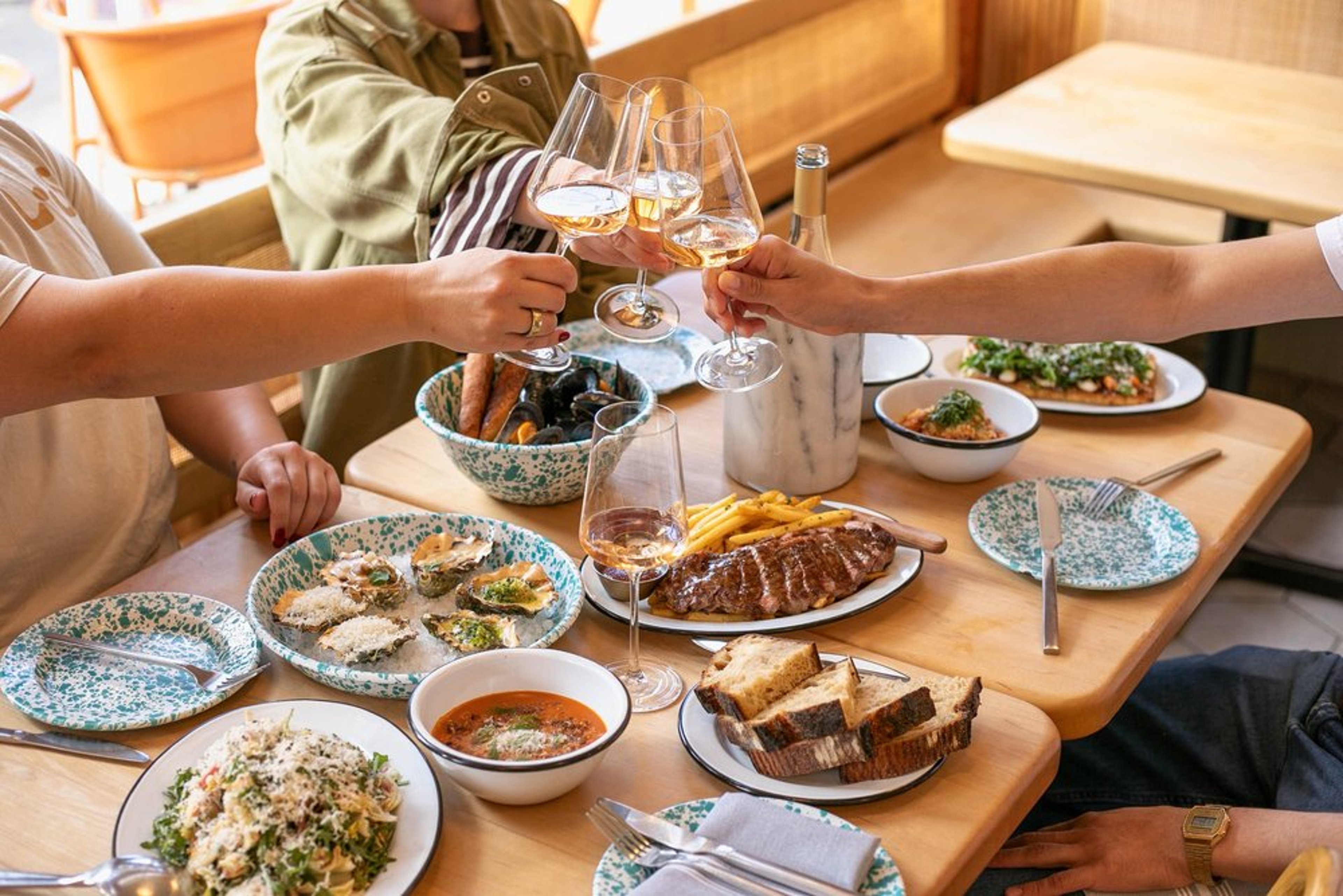 People celebrating with clinking glasses of rosé wine at a dinner table with various dishes including oysters, steak, pasta, bread, and bowls of soup and vegetables.
