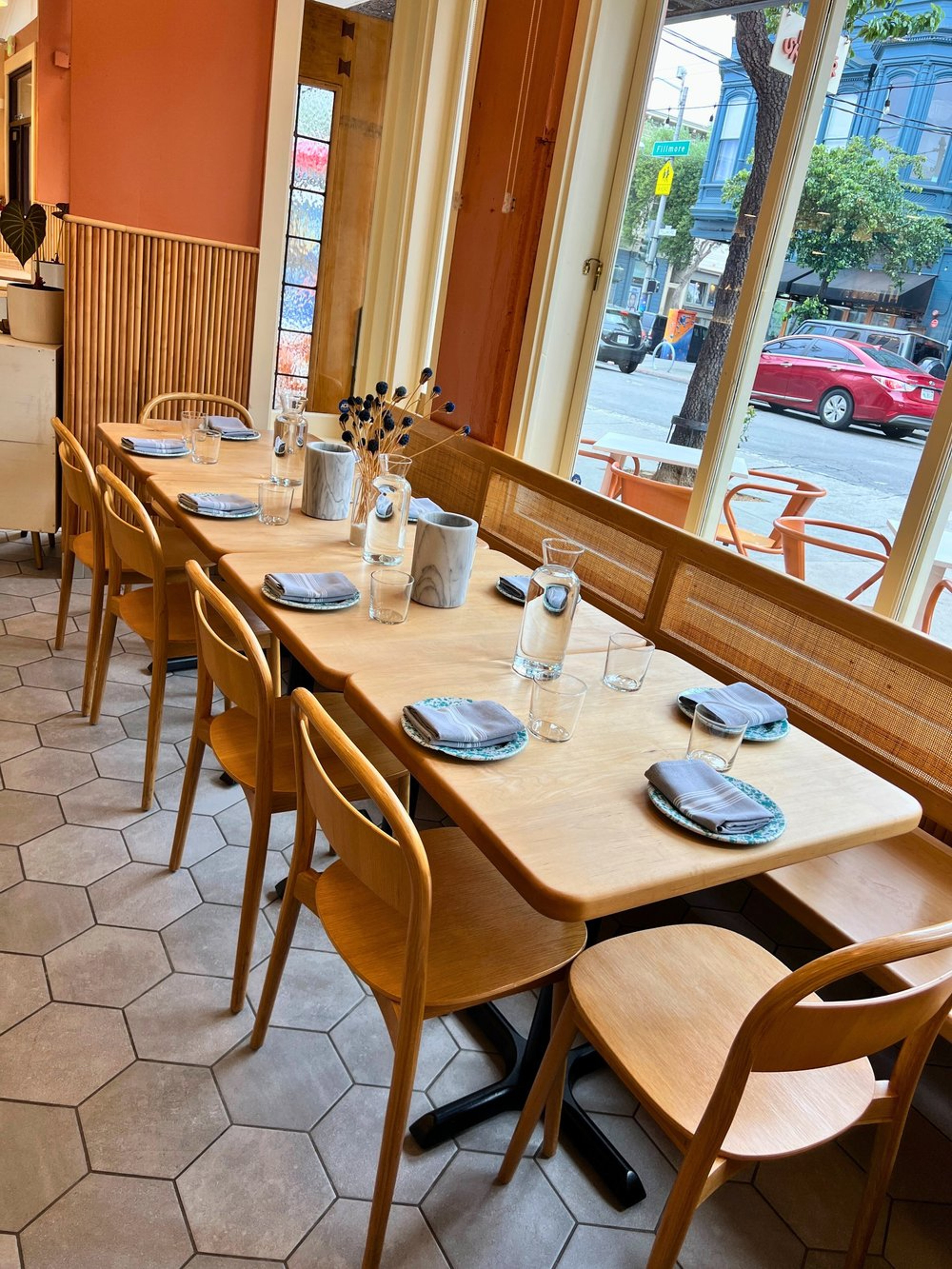 A cozy restaurant interior with a long wooden table set for dining, including glasses, napkins, and decorative vases with dried flowers, next to large windows showing a city street with parked cars and buildings outside.